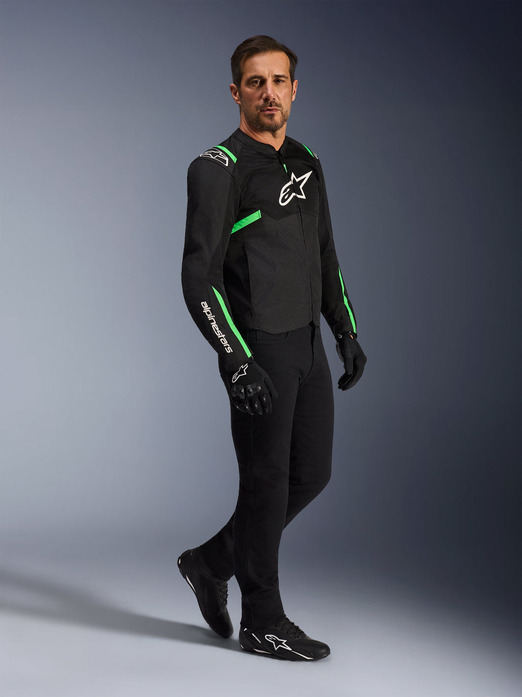 Męski model w kurtce Alpinestars T-SPS Air V2 w kolorze czarnym i Fluo Green, w zestawieniu z czarnymi spodniami i rękawicami motocyklowymi, sportowa kurtka tekstylna z panelami mesh i neonowymi akcentami, kompletny miejski strój motocyklowy
