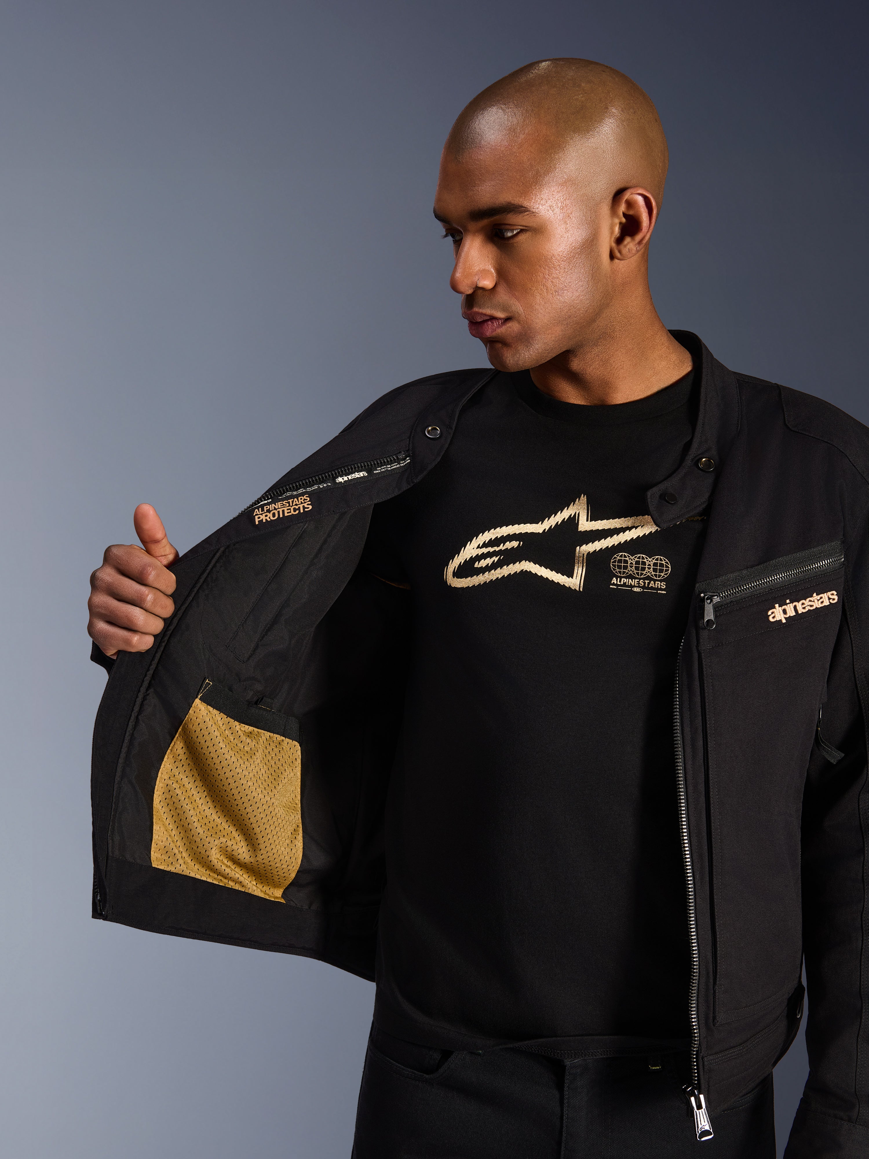 Alpinestars Bruiser Jacket, czarna miejska kurtka motocyklowa, zbliżenie na otwarte wnętrze ze złotą kieszenią z siateczki i napisem „Alpinestars Protects”, założona na czarny t-shirt z logo, tekstylna odzież motocyklowa o estetyce odzieży roboczej