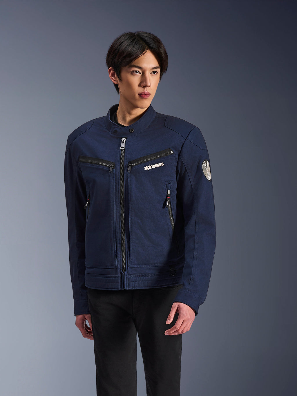 Alpinestars Bruiser Jacket, granatowa tekstylna kurtka motocyklowa, noszona przez mężczyznę w czarnych spodniach, z kieszeniami na zamek, białym logo Alpinestars na piersi i okrągłą spiralną naszywką Oscar na rękawie, miejski styl streetwear dla motocyklistów
