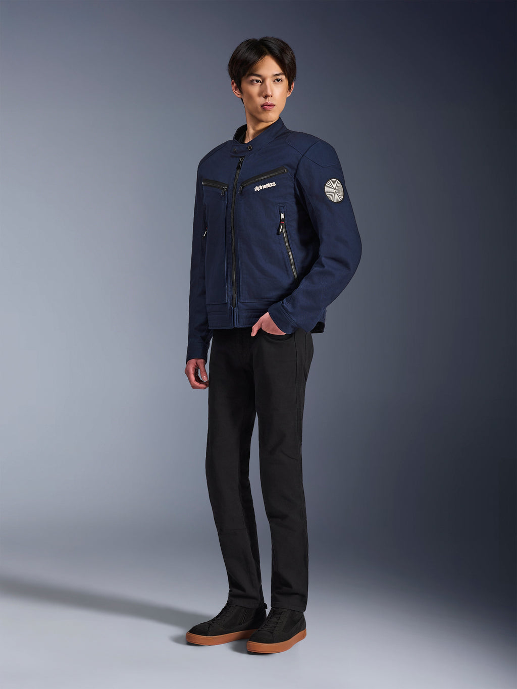Alpinestars Bruiser Jacket w kolorze Dark Blue, miejska tekstylna kurtka motocyklowa o designie inspirowanym odzieżą roboczą, z zapinanymi na zamek kieszeniami na piersi i spiralną naszywką Oscar na rękawie, noszona z czarnymi spodniami i codziennymi sneakersami.