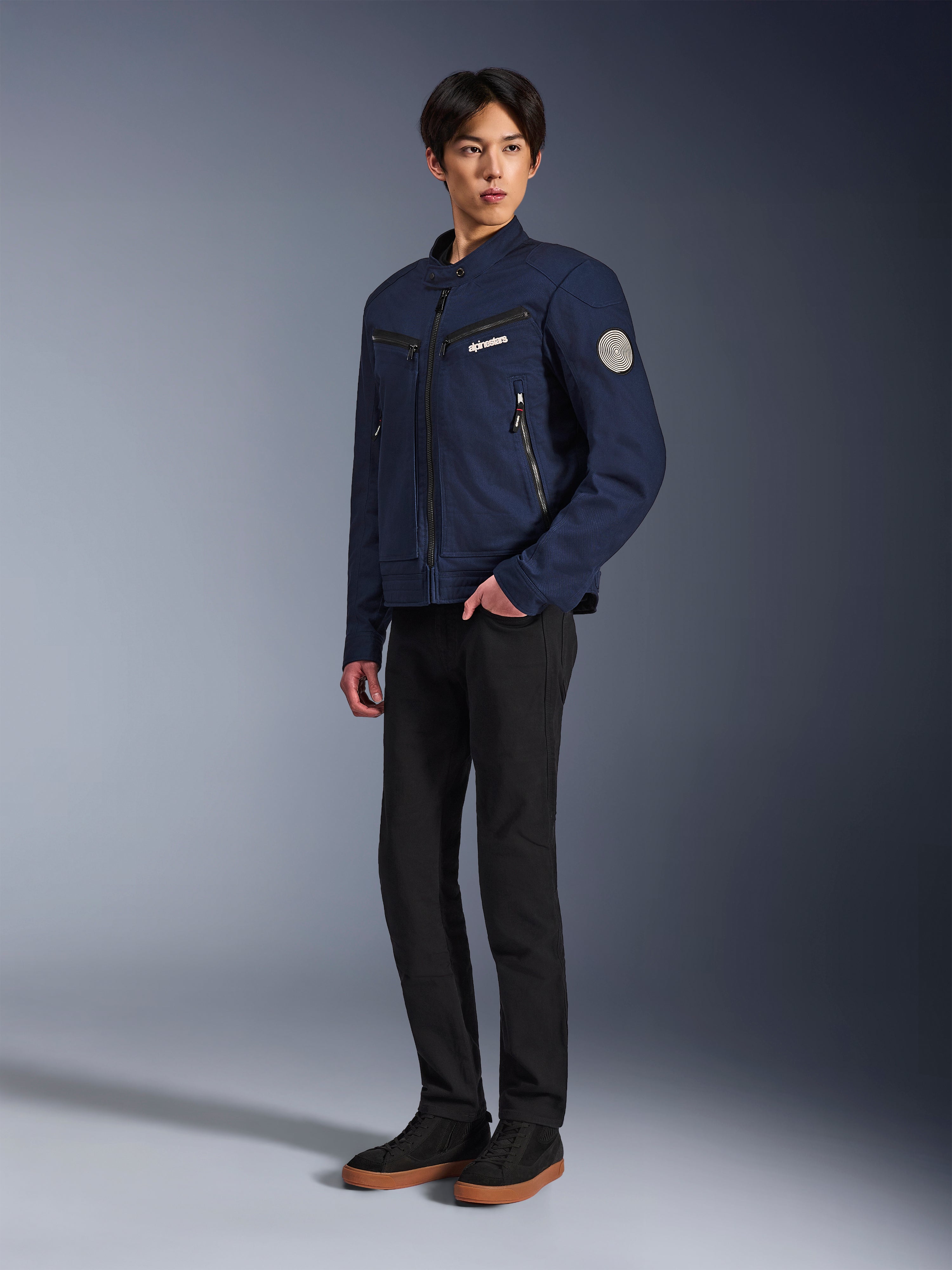 Alpinestars Bruiser Jacket w kolorze Dark Blue, miejska tekstylna kurtka motocyklowa o designie inspirowanym odzieżą roboczą, z zapinanymi na zamek kieszeniami na piersi i spiralną naszywką Oscar na rękawie, noszona z czarnymi spodniami i codziennymi sneakersami.