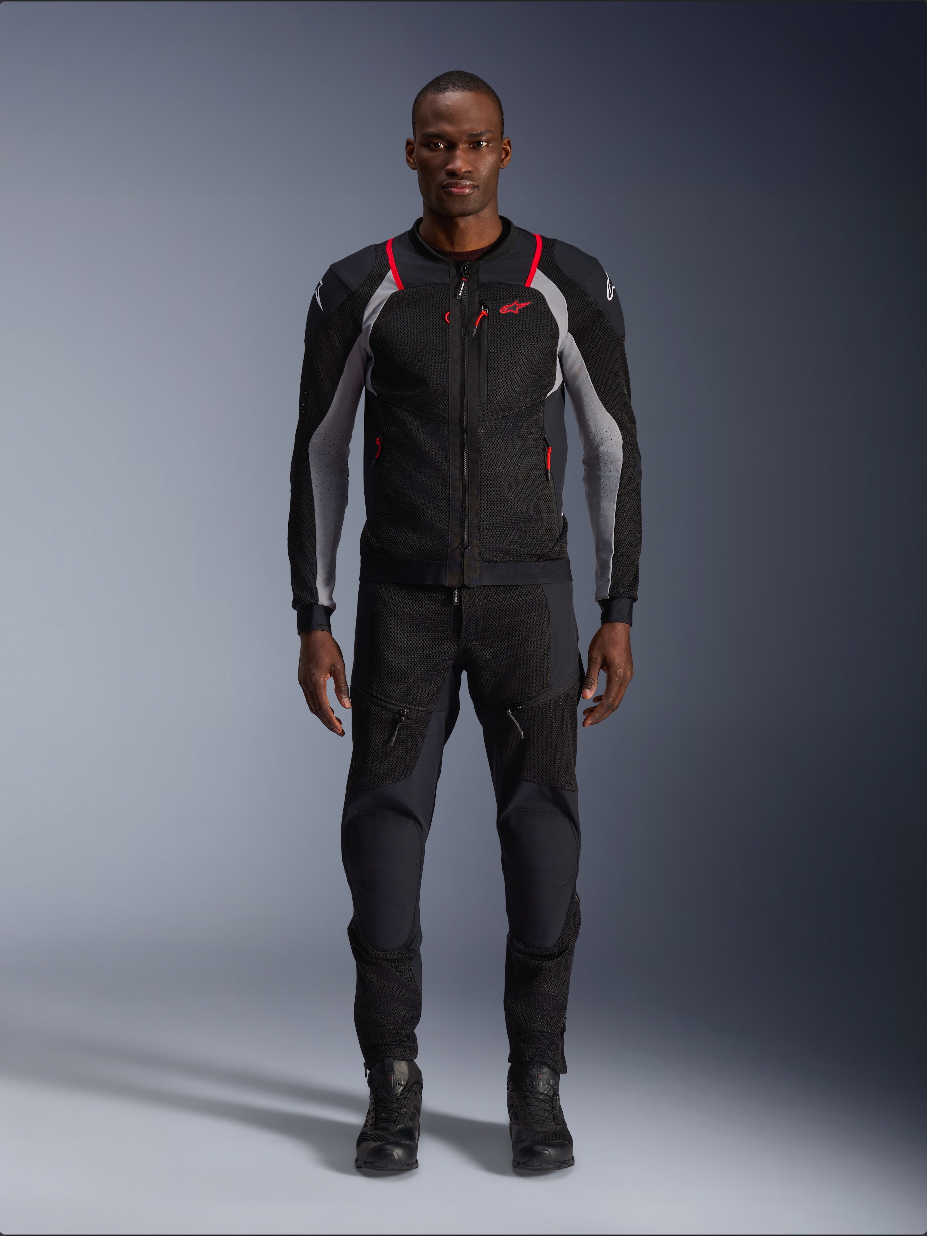 Alpinestars Troop-Air Pro Jacket, techniczna kurtka motocyklowa, czarna, popielato-szara i jaskrawoczerwona, z panelami z siatki i czerwonymi akcentami, noszona przez modela z czarnymi spodniami i butami motocyklowymi, nowoczesny miejski design z techniczną wentylacją.