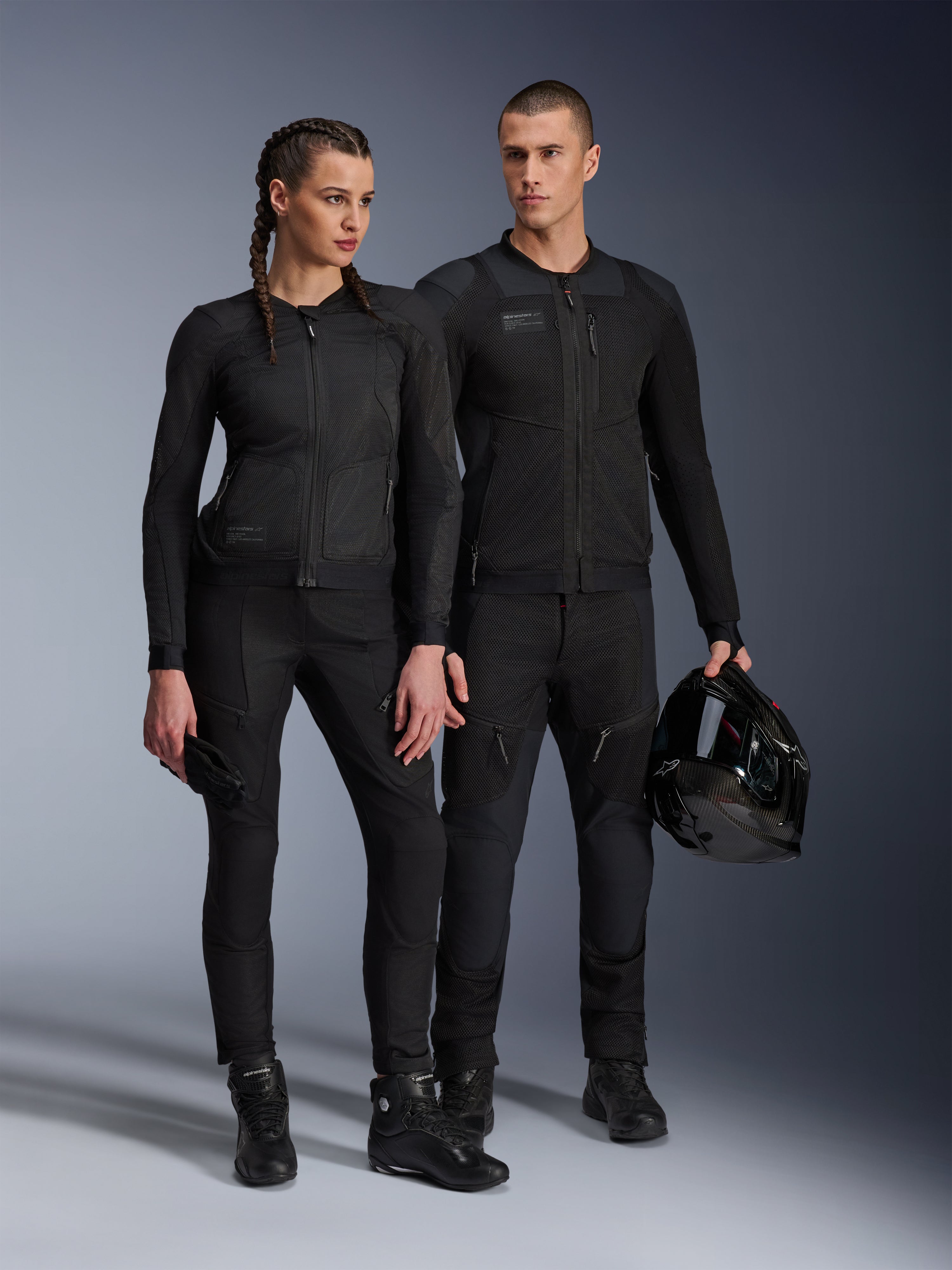 Kurtka Alpinestars Stella Troop-Air, mężczyzna i kobieta w pasującej do siebie, całkowicie czarnej technicznej odzieży motocyklowej z siatkową konstrukcją zapewniającą przepływ powietrza, czarne spodnie i buty, mężczyzna trzymający czarny kask, miejska odzież motocyklowa