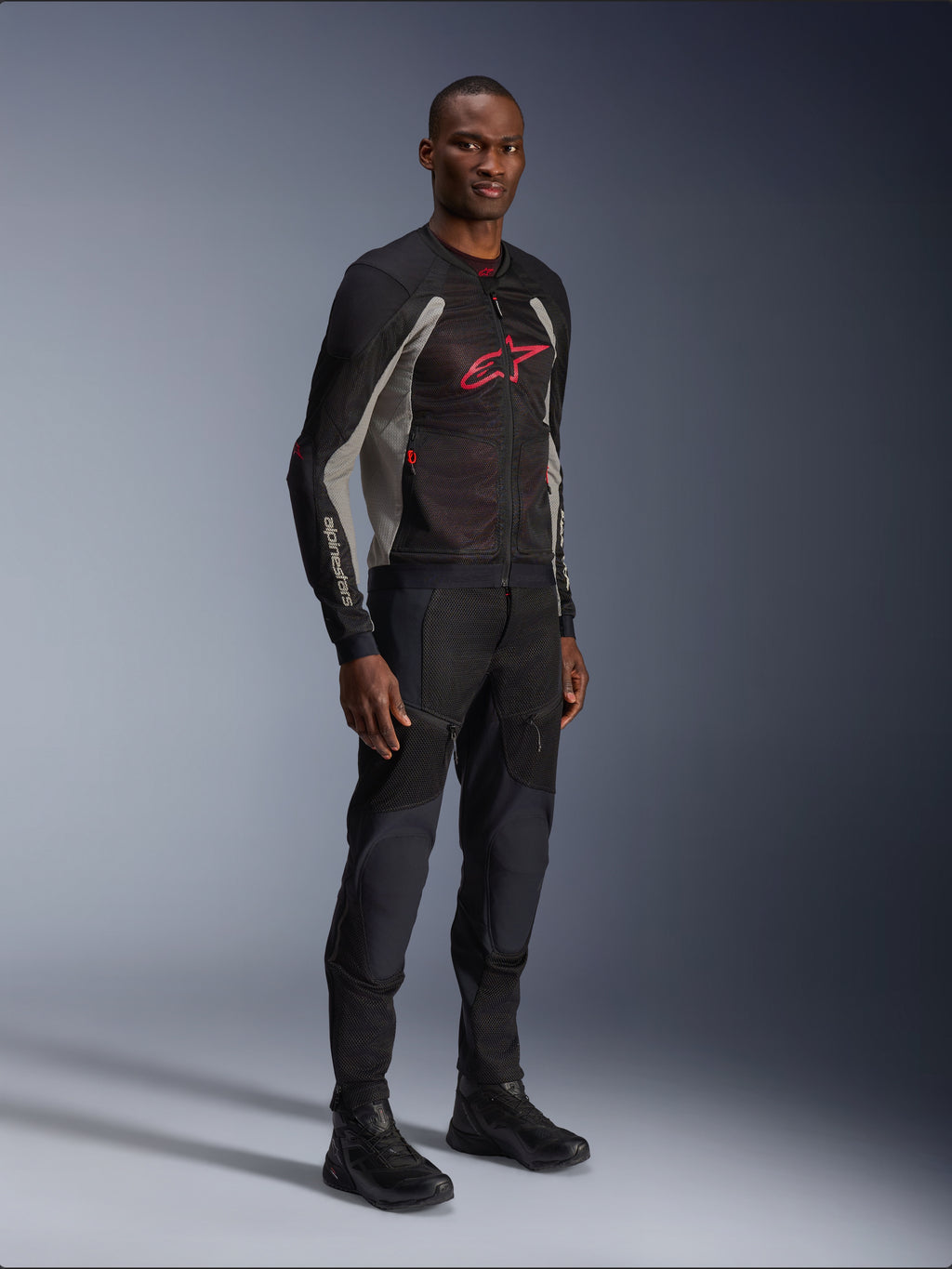 Kurtka Alpinestars Troop-Air, techniczna siatkowa kurtka motocyklowa w kolorach czarnym, popielatym i jasnoczerwonym, prezentowana przez modela z czarnymi spodniami i butami, czerwone logo Alpinestars na piersi, nowoczesna odzież motocyklowa w stylu miejskim.
