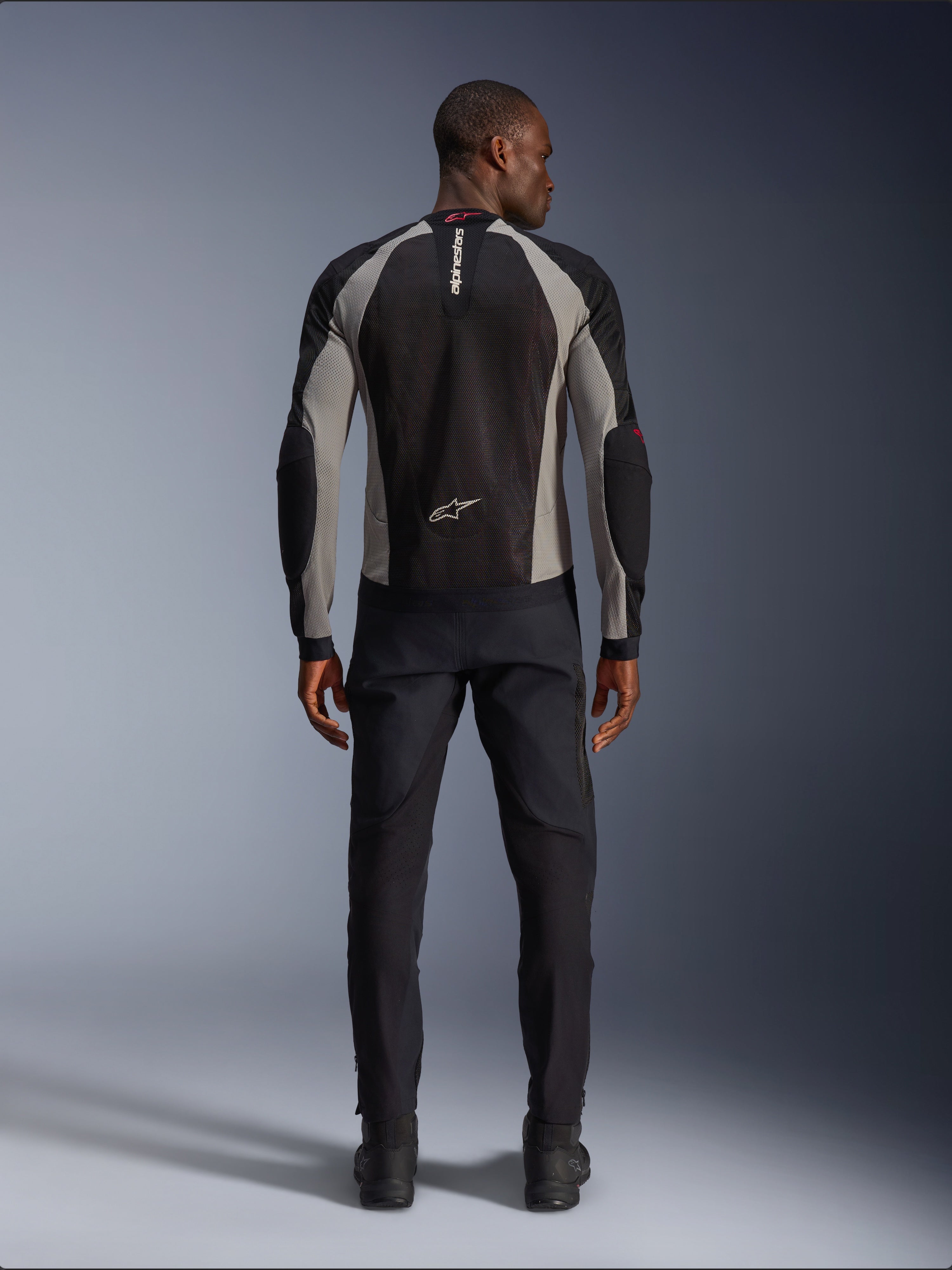 Alpinestars Troop-Air Jacket, motocyklowa kurtka w kolorze czarnym, popielatym i jaskrawoczerwonym, widok z tyłu na modelu, lekka siatkowa konstrukcja zapewniająca wentylację, branding Alpinestars na kołnierzu i w dolnej części pleców, w zestawieniu z czarnymi spodniami motocyklowymi