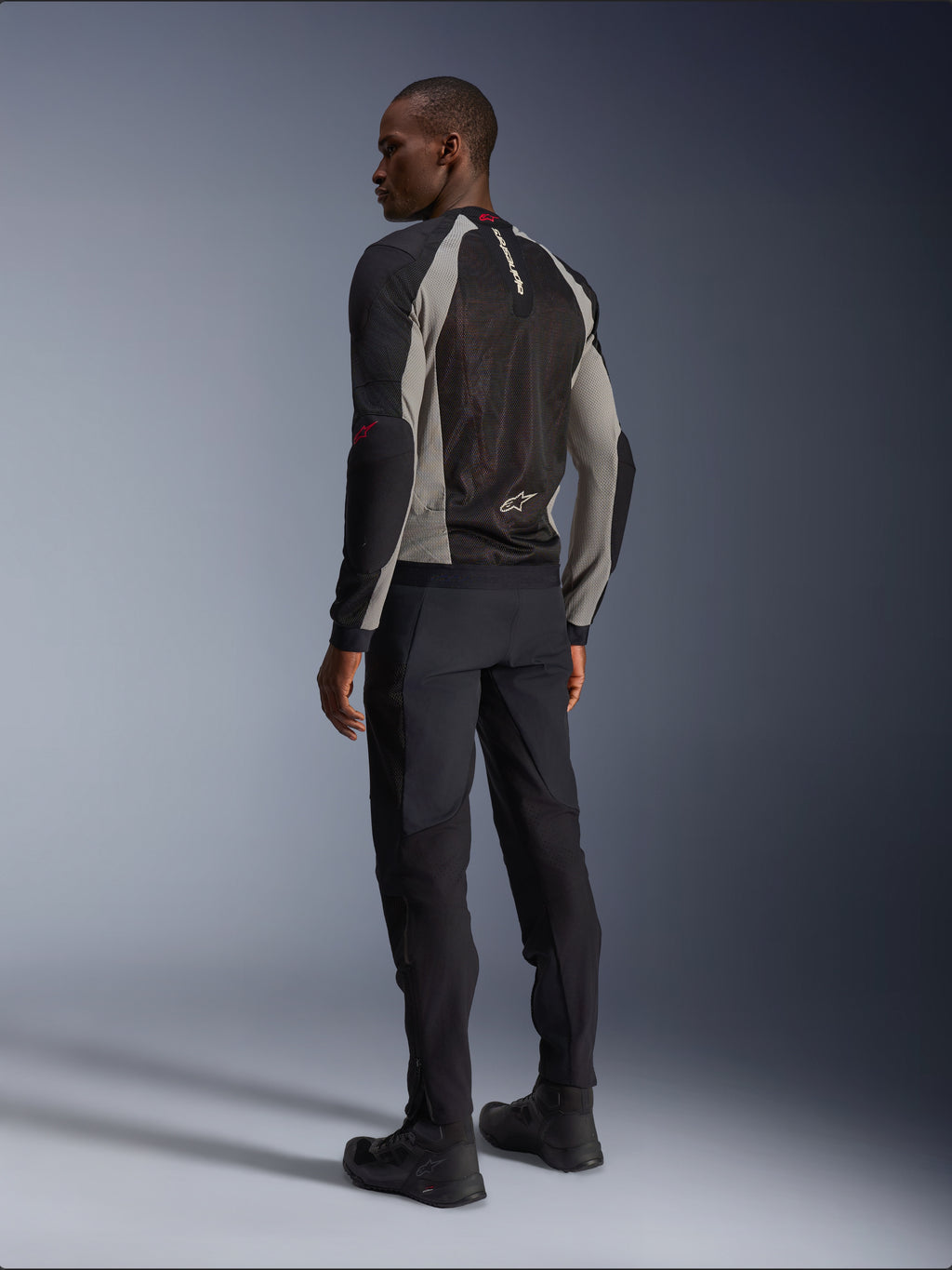 Model w kurtce Alpinestars Troop-Air, widok od tyłu, kolorystyka Black Ash Gray z jaskrawoczerwonymi akcentami, techniczna konstrukcja z siatki mesh dla optymalnej wentylacji, czarne spodnie motocyklowe i buty, współczesny styl miejski