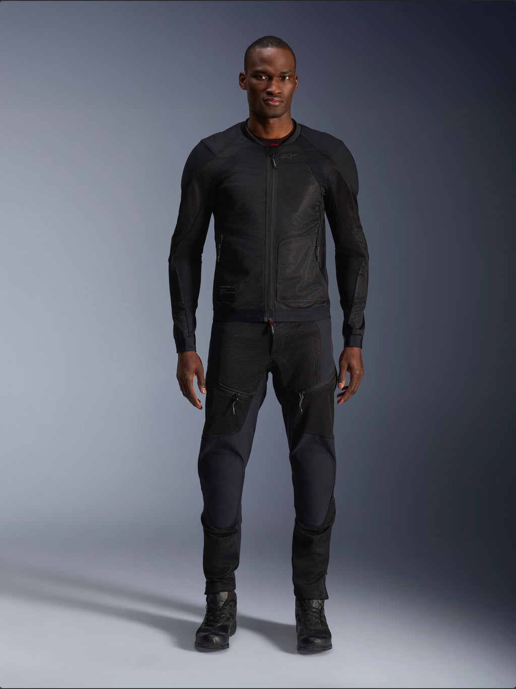Męski model w kurtce Alpinestars Troop-Air Jacket w kolorze Black Black, lekka siatkowa kurtka motocyklowa, połączona z dopasowanymi czarnymi spodniami i butami motocyklowymi, techniczna konstrukcja z siatki 2D zapewniająca doskonały przepływ powietrza.