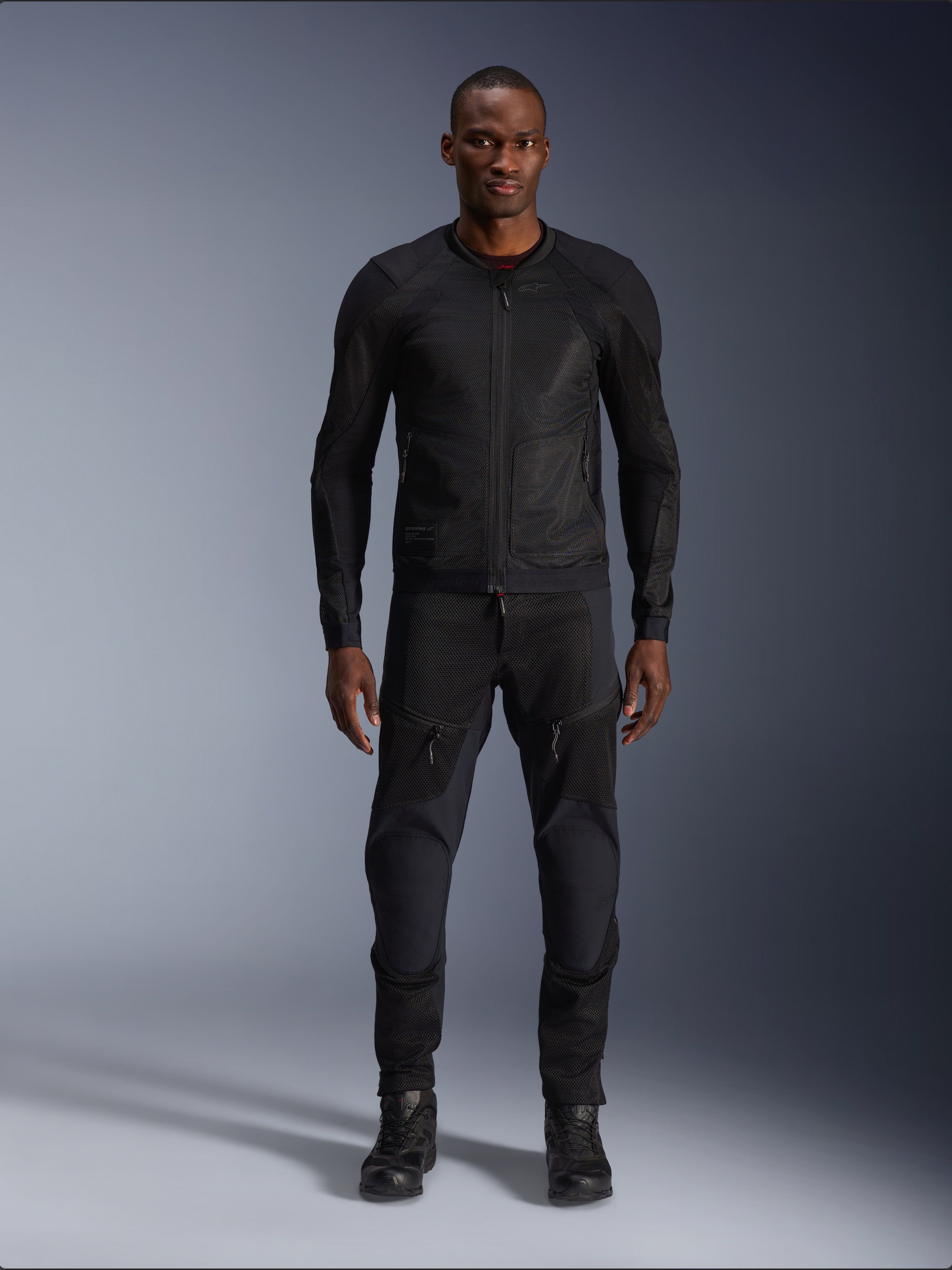 Męski model w kurtce Alpinestars Troop-Air Jacket w kolorze Black Black, lekka siatkowa kurtka motocyklowa, połączona z dopasowanymi czarnymi spodniami i butami motocyklowymi, techniczna konstrukcja z siatki 2D zapewniająca doskonały przepływ powietrza.