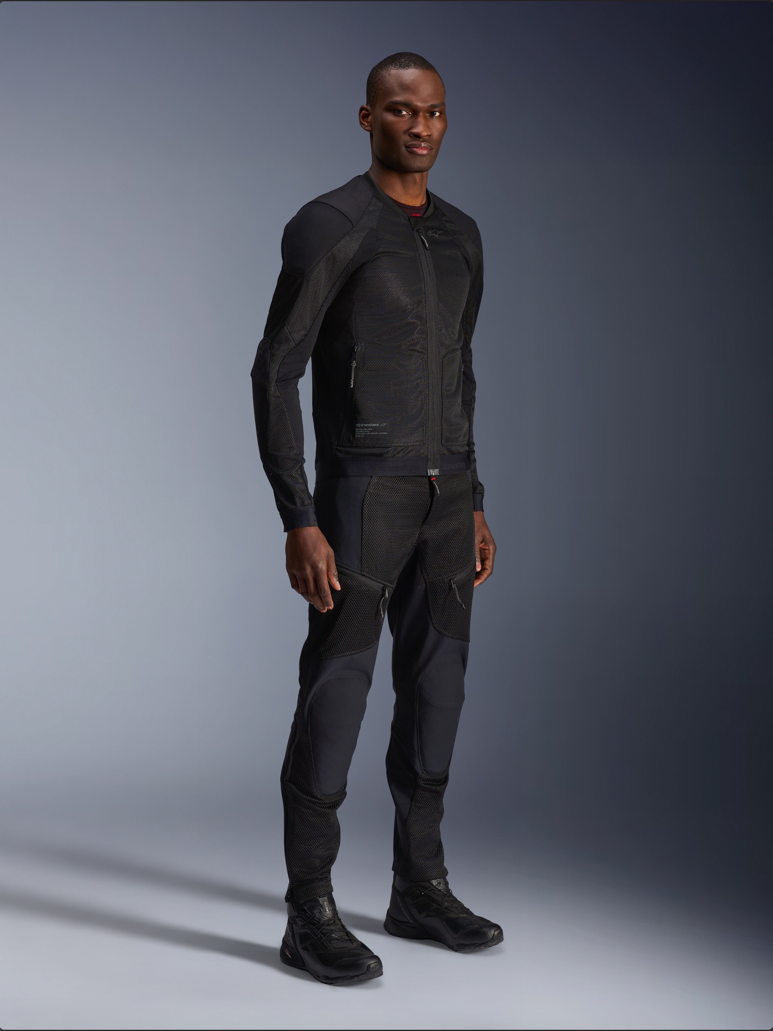Alpinestars Troop-Air Jacket, czarna, techniczna siatkowa kurtka motocyklowa prezentowana przez modela z pasującymi czarnymi spodniami i butami, lekka konstrukcja z siatki i minimalistyczny miejski design