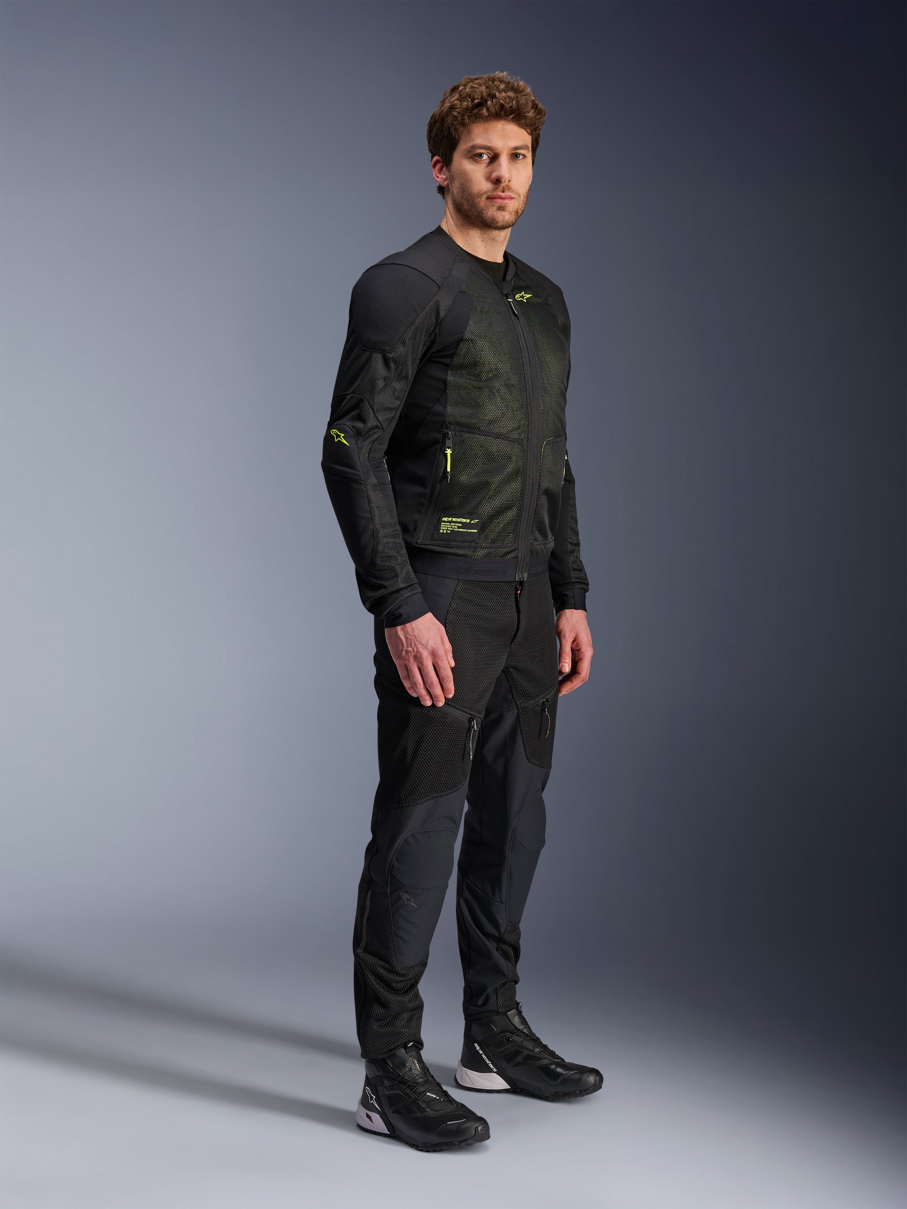 Kurtka Alpinestars Troop-Air, motocyklowa kurtka z siatki, czarno-żółta fluo, noszona przez modela z dopasowanymi czarnymi spodniami i butami motocyklowymi, posiada konstrukcję z siatki o wysokim przepływie powietrza z żółtymi fluorescencyjnymi akcentami na klatce piersiowej i rękawach, lekki techniczny design do jazdy szosowej