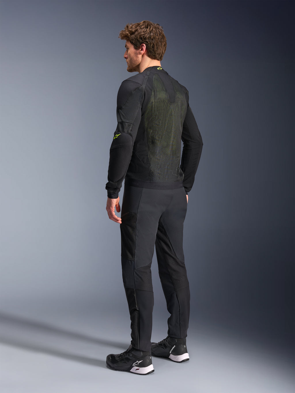 Kurtka Alpinestars Troop-Air w kolorze czarnym i żółtym fluo, widok z tyłu na męskiego modela ubranego w techniczną kurtkę powietrzną, z dużym tylnym panelem z siatki z żółtymi akcentami i fluorescencyjnymi żółtymi logo Alpinestars, w zestawieniu z czarnymi spodniami motocyklowymi.