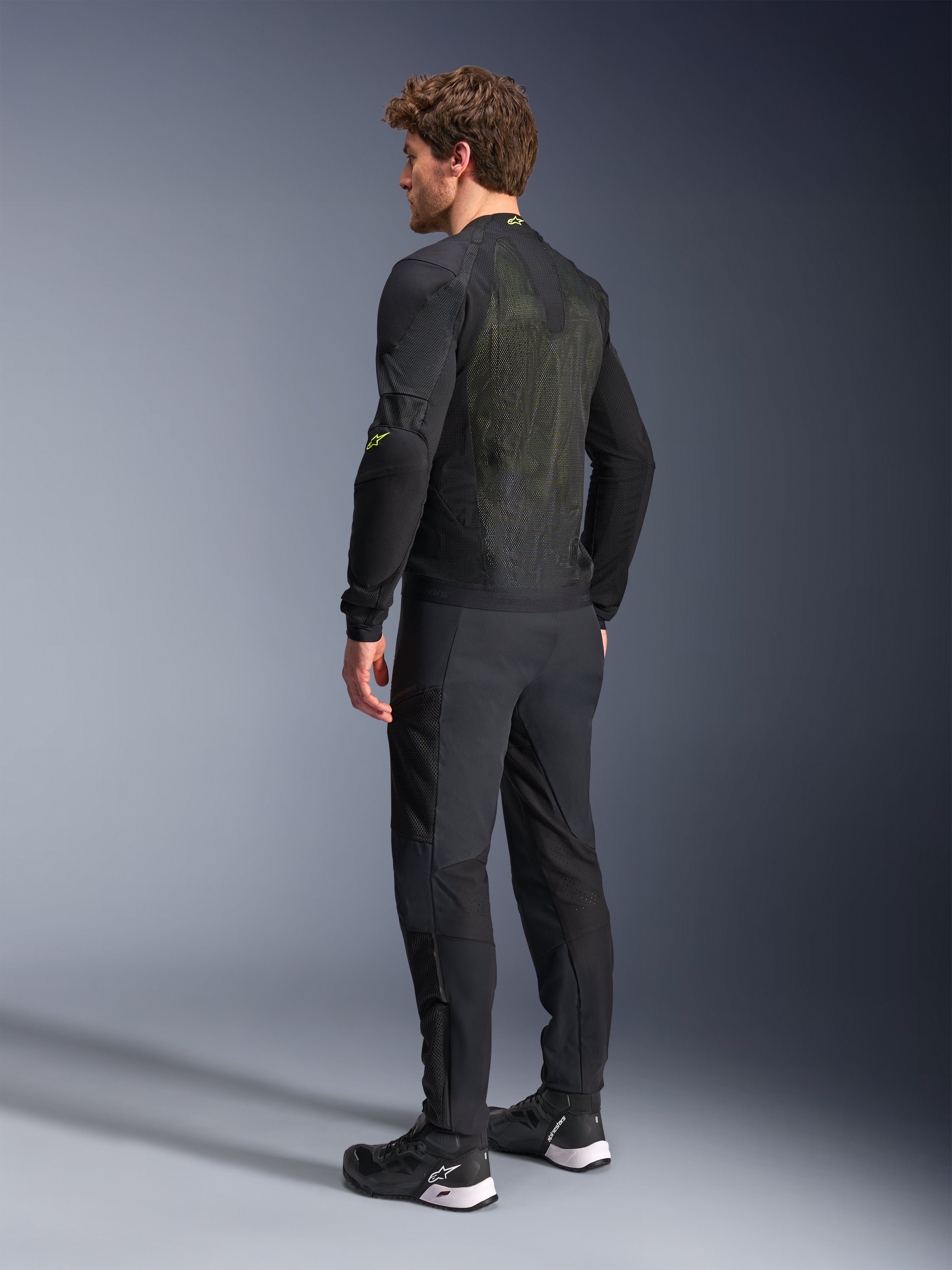 Kurtka Alpinestars Troop-Air w kolorze czarnym i żółtym fluo, widok z tyłu na męskiego modela ubranego w techniczną kurtkę powietrzną, z dużym tylnym panelem z siatki z żółtymi akcentami i fluorescencyjnymi żółtymi logo Alpinestars, w zestawieniu z czarnymi spodniami motocyklowymi.