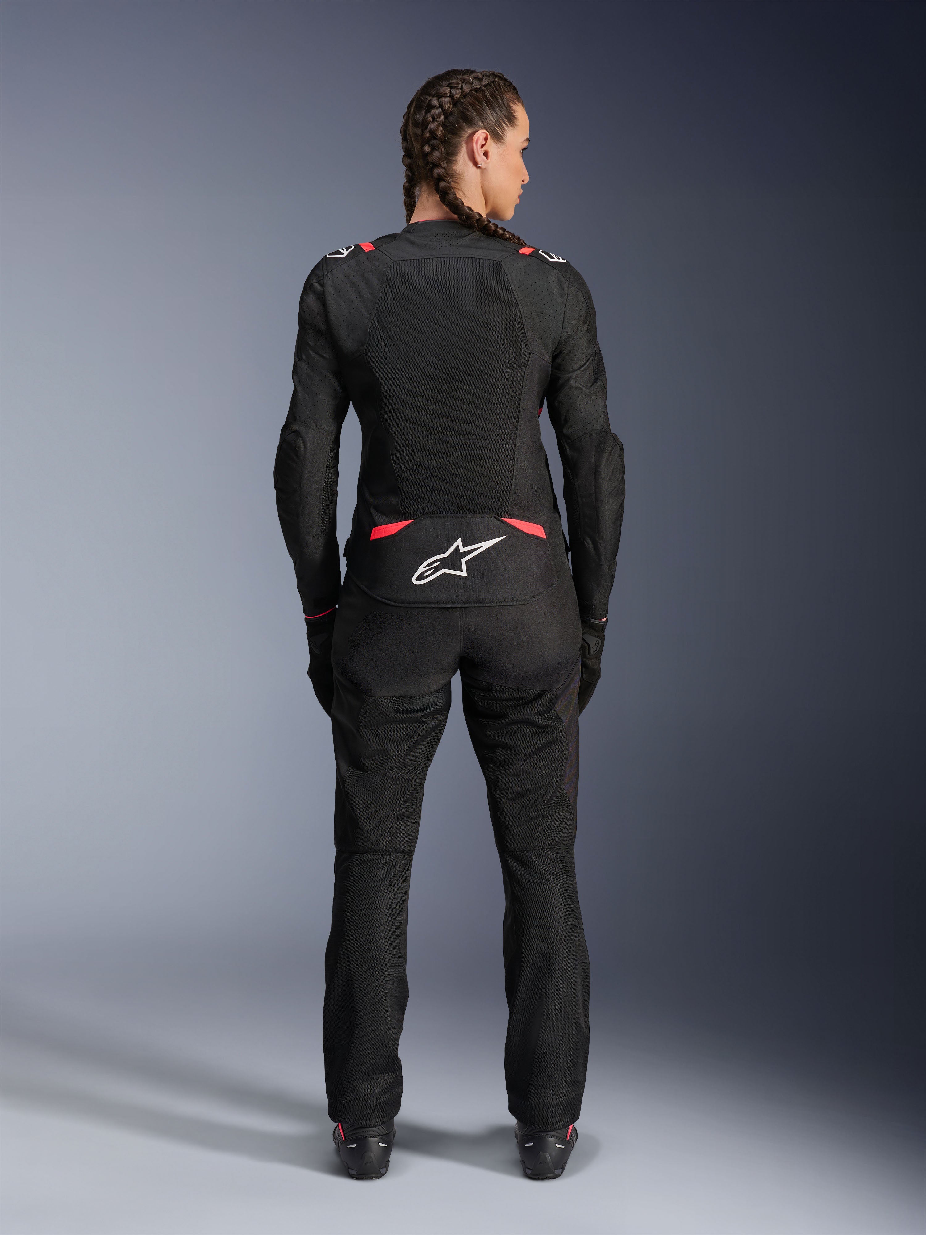 Alpinestars Stella T-SPS Air V2 Jacket, damska tekstylna kurtka motocyklowa, czarna z różowymi akcentami Diva Pink, widok z tyłu na modelce z warkoczami, duże panele z siatki dla wentylacji, białe logo Astars w dolnej części pleców, w zestawie z dopasowanymi czarnymi spodniami motocyklowymi.