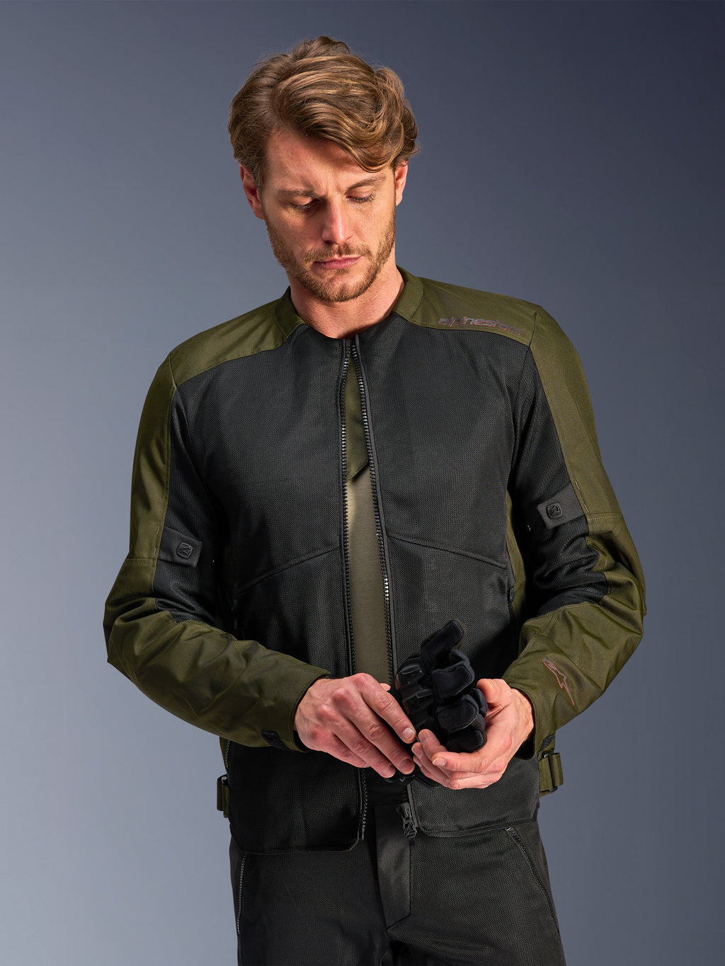 Man wearing the Alpinestars C-1 Air Jacket in Khaki, urban motorcycle jacket with olive green textile and black mesh panels, looking down while holding black motorcycle gloves, clean and contemporary style for road riding.