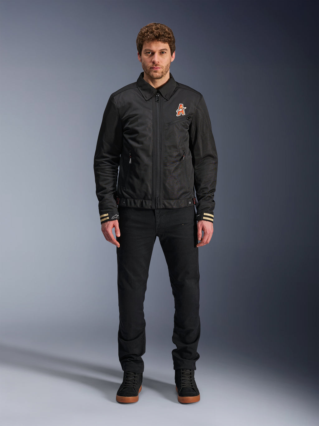 Męski model w czarnej kurtce Alpinestars Ignite Air z klasycznym kołnierzykiem koszulowym, pełnym zamkiem błyskawicznym z przodu i pomarańczowym logo na piersi. Stylizacja z czarnymi spodniami i czarnymi sneakersami za kostkę tworzy współczesny miejski wygląd motocyklowy.
