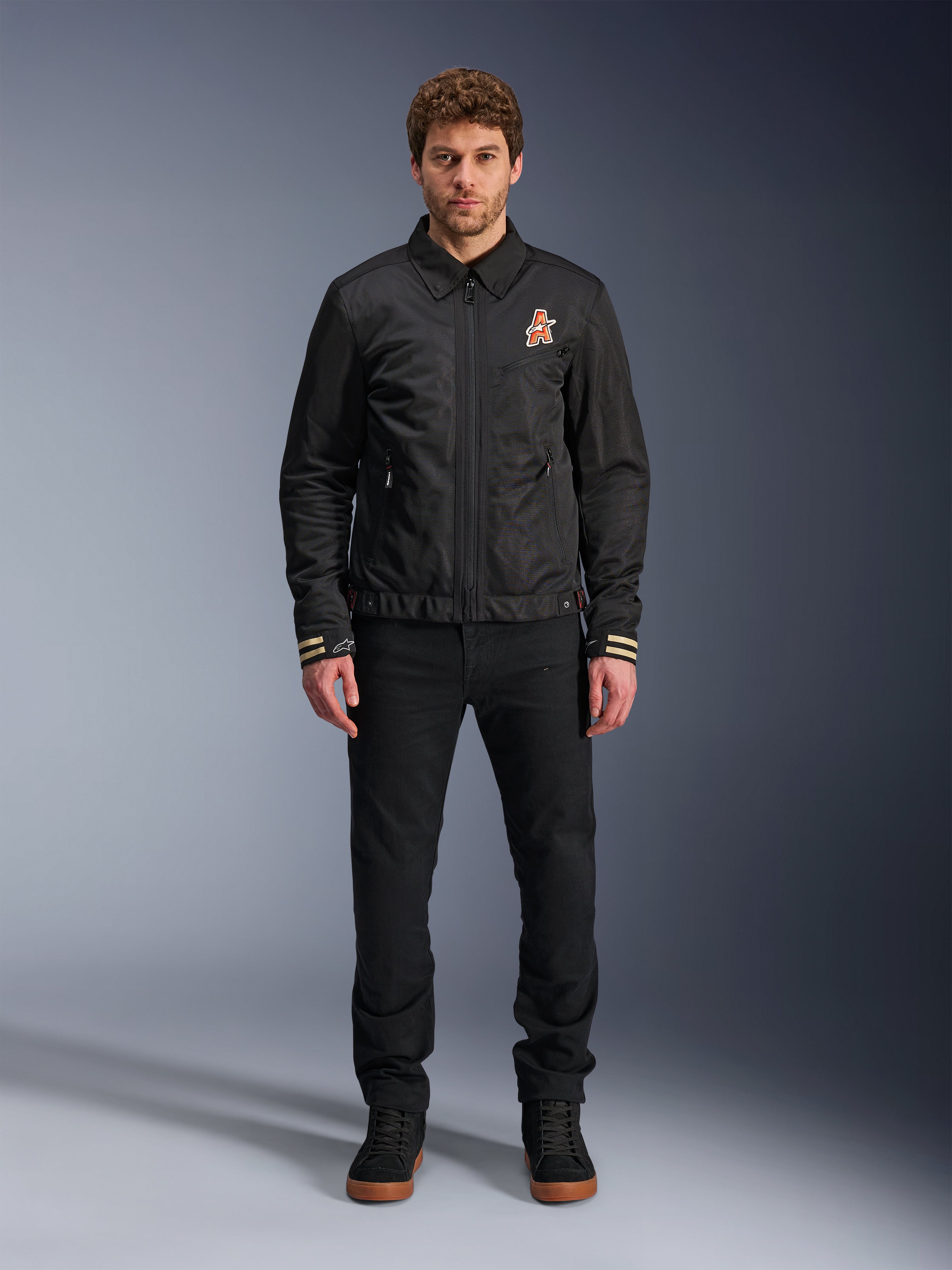 Męski model w czarnej kurtce Alpinestars Ignite Air z klasycznym kołnierzykiem koszulowym, pełnym zamkiem błyskawicznym z przodu i pomarańczowym logo na piersi. Stylizacja z czarnymi spodniami i czarnymi sneakersami za kostkę tworzy współczesny miejski wygląd motocyklowy.