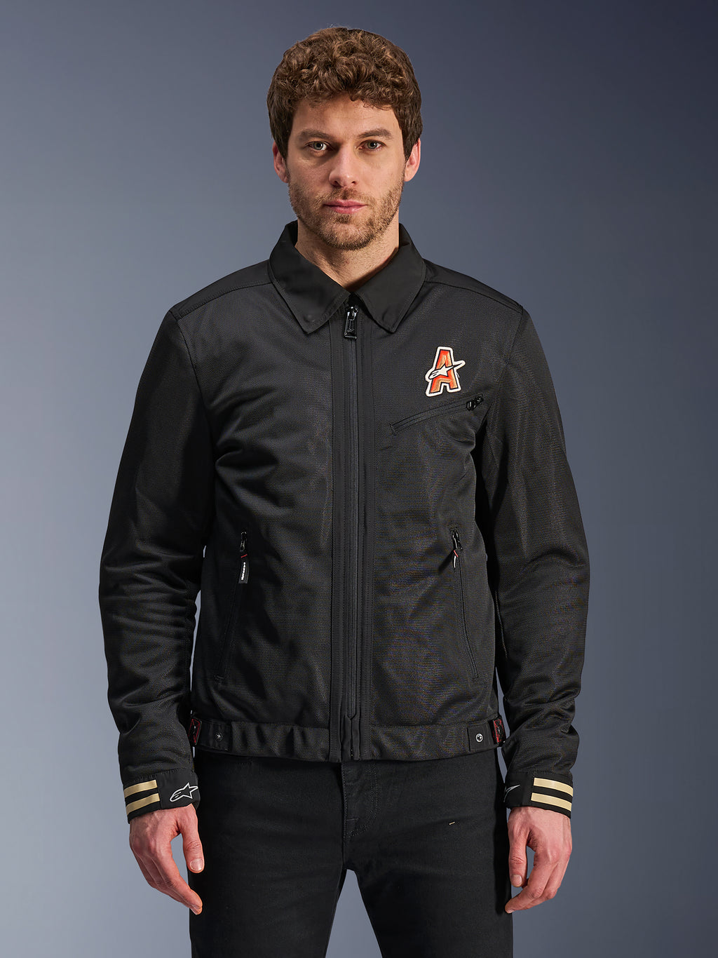 Alpinestars Ignite Air Jacket, czarna kurtka motocyklowa, tradycyjny kołnierzyk koszulowy, pomarańczowo-białe logo „A” na piersi, mankiety w beżowe paski z białym logo gwiazdy, nowoczesna miejska odzież motocyklowa