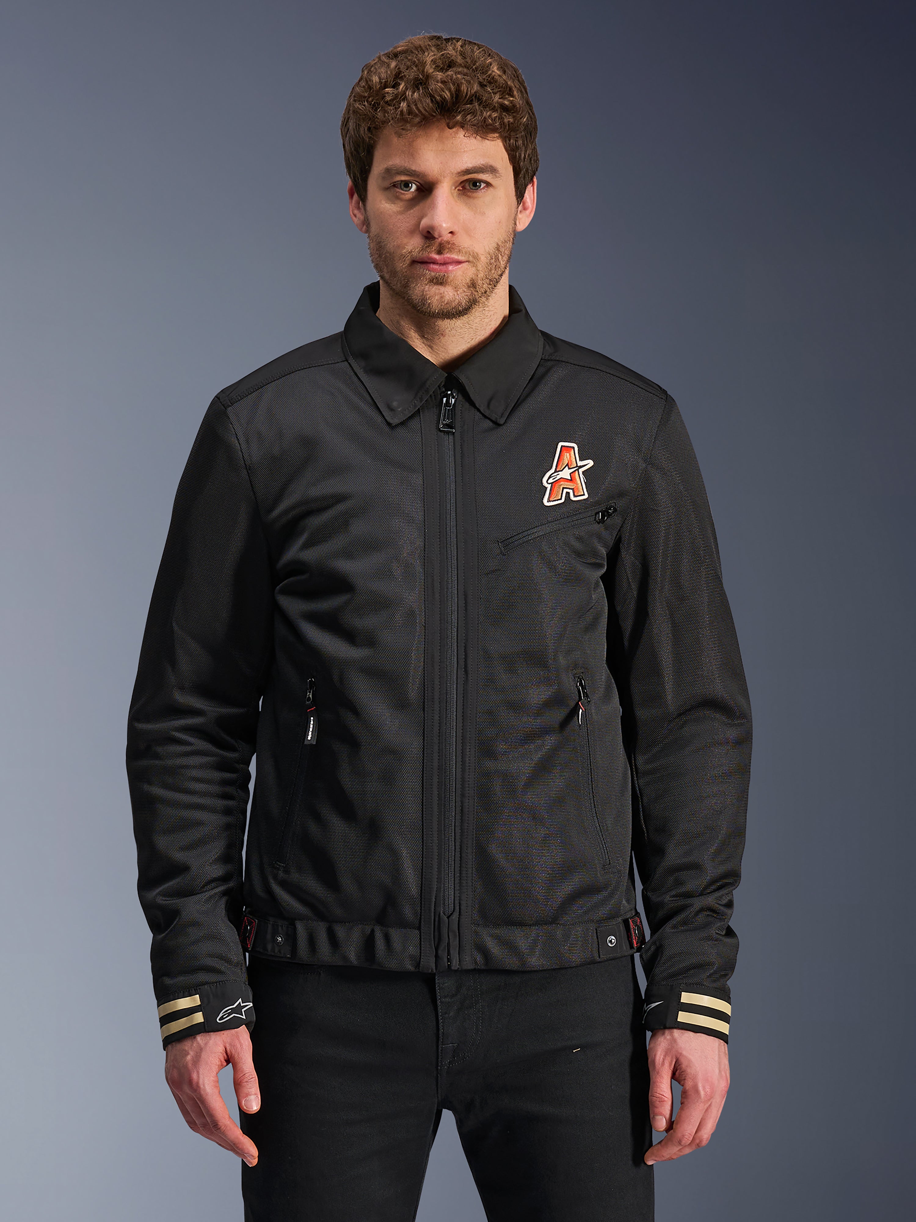 Alpinestars Ignite Air Jacket, czarna kurtka motocyklowa, tradycyjny kołnierzyk koszulowy, pomarańczowo-białe logo „A” na piersi, mankiety w beżowe paski z białym logo gwiazdy, nowoczesna miejska odzież motocyklowa