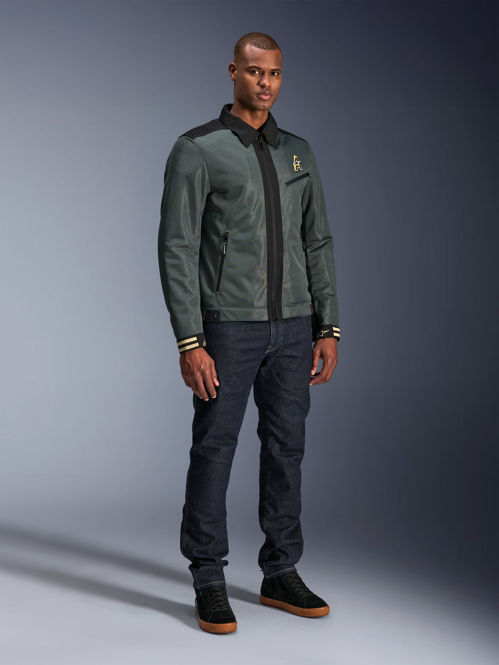 Alpinestars Ignite Air Jacket, Urban Gray Black Blue, miejska kurtka motocyklowa, noszona przez modela z ciemnymi jeansami i czarnymi sneakersami, siateczkowy korpus, tradycyjny kołnierzyk koszulowy, fason inspirowany kurtką Harrington i złote logo Alpinestars.