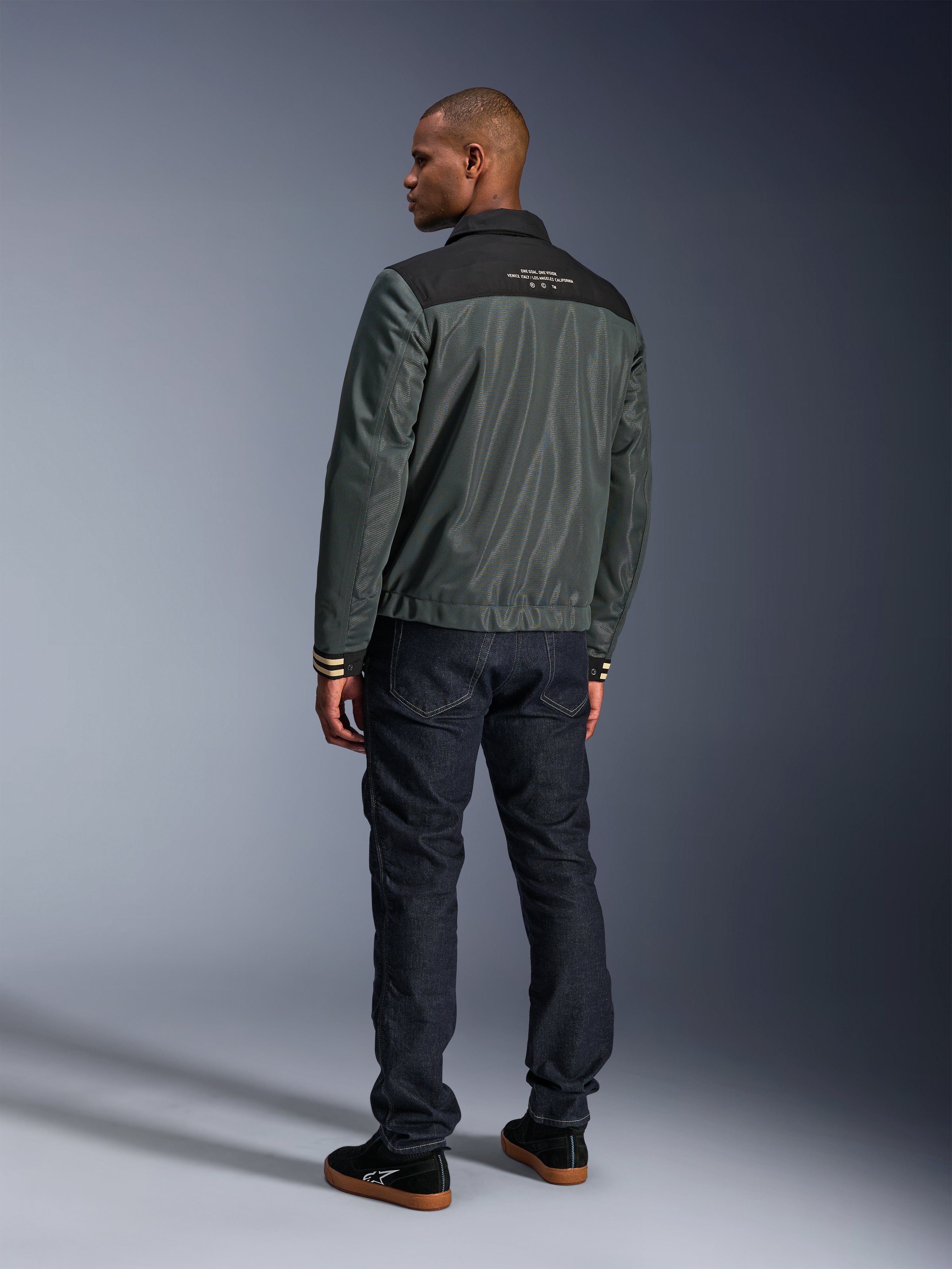 Kurtka Alpinestars Ignite Air w kolorze Urban Gray Black Blue, widok z tyłu na modelu, siatkowy korpus z czarnym karczkiem i białym napisem, prążkowane mankiety, w zestawieniu z ciemnymi jeansami i czarnymi sneakersami, miejska odzież motocyklowa lifestyle