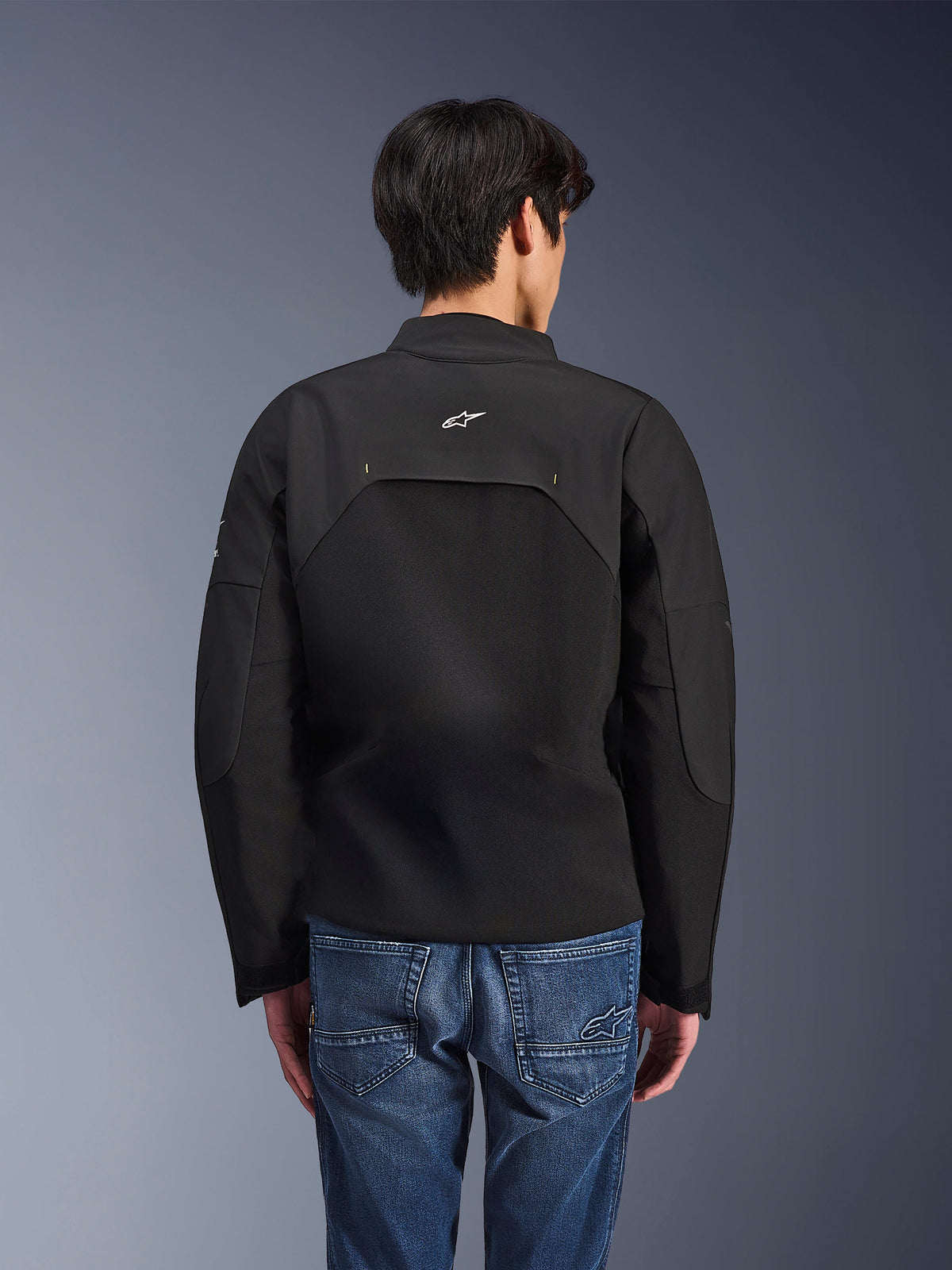 Kurtka Alpinestars Provoke, miejska kurtka motocyklowa, czarna, widok z tyłu przedstawiający białe logo Astars i techniczne panele ripstop, noszona z niebieskimi jeansami, nowoczesny design do miejskich dojazdów