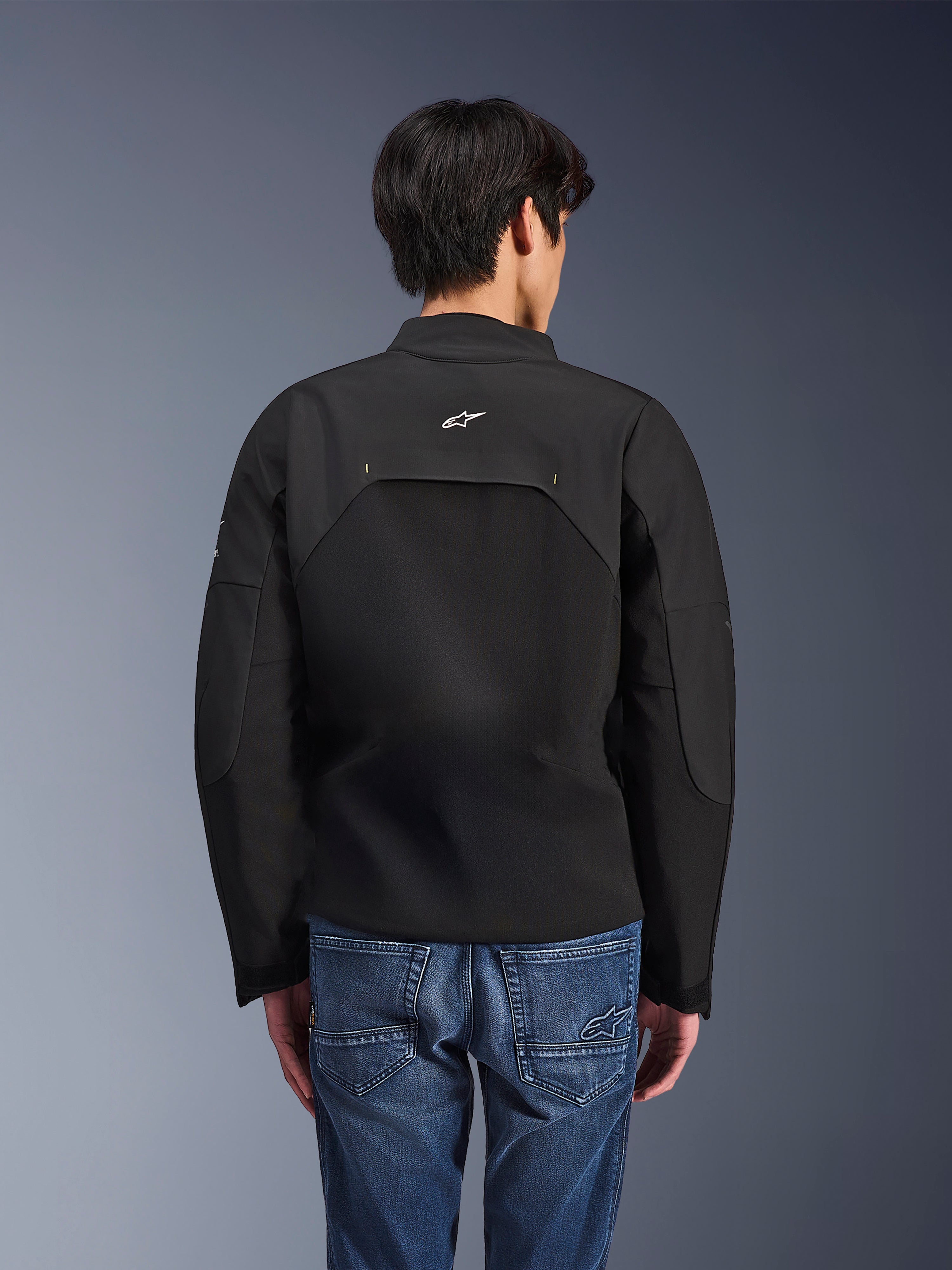Kurtka Alpinestars Provoke, miejska kurtka motocyklowa, czarna, widok z tyłu przedstawiający białe logo Astars i techniczne panele ripstop, noszona z niebieskimi jeansami, nowoczesny design do miejskich dojazdów
