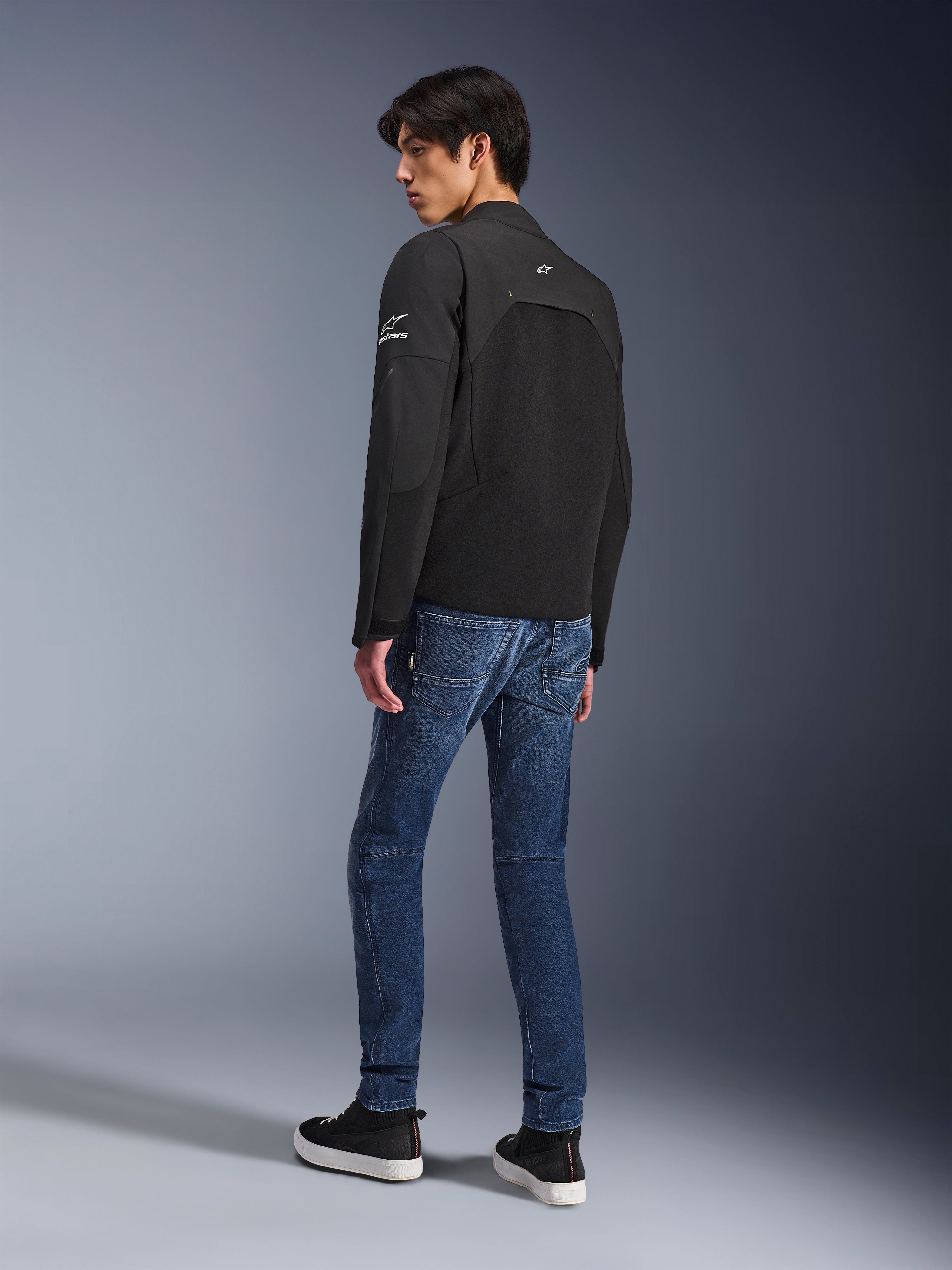 Kurtka Alpinestars Provoke, czarna miejska kurtka motocyklowa, model w ujęciu z tyłu pod kątem trzech czwartych, zestawiona z niebieskimi jeansami i czarnymi sneakersami, wstawki softshell ripstop oraz białe logo Alpinestars na rękawie i plecach.