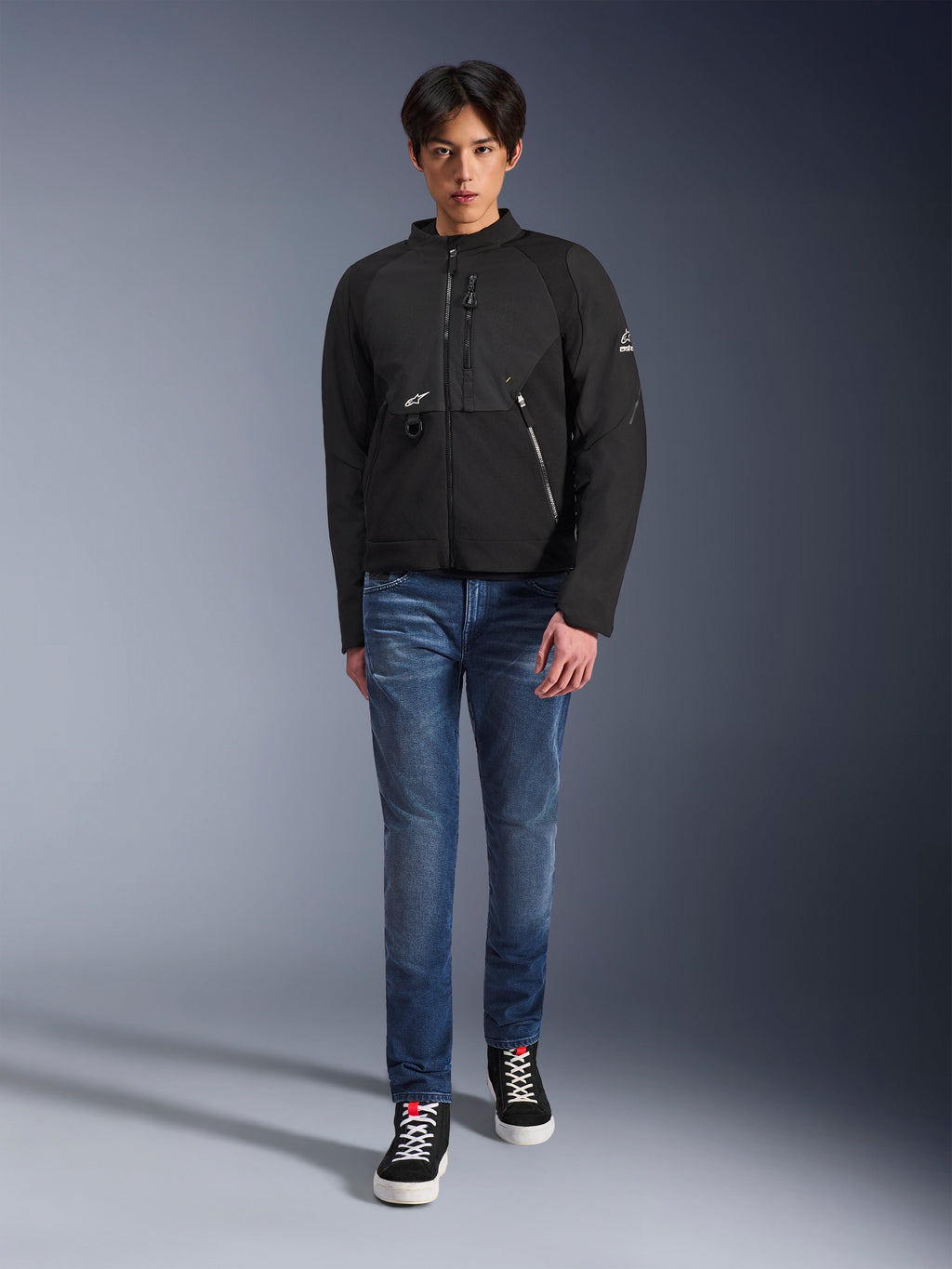 Męski model w czarnej kurtce Alpinestars Provoke, miejska tekstylna kurtka motocyklowa z technicznymi zamkami i panelami softshell ripstop, zestawiona z niebieskimi dżinsami i czarnymi sneakersami, współczesny miejski styl.