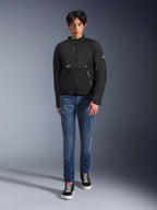 Męski model w czarnej kurtce Alpinestars Provoke, miejska tekstylna kurtka motocyklowa z technicznymi zamkami i panelami softshell ripstop, zestawiona z niebieskimi dżinsami i czarnymi sneakersami, współczesny miejski styl.