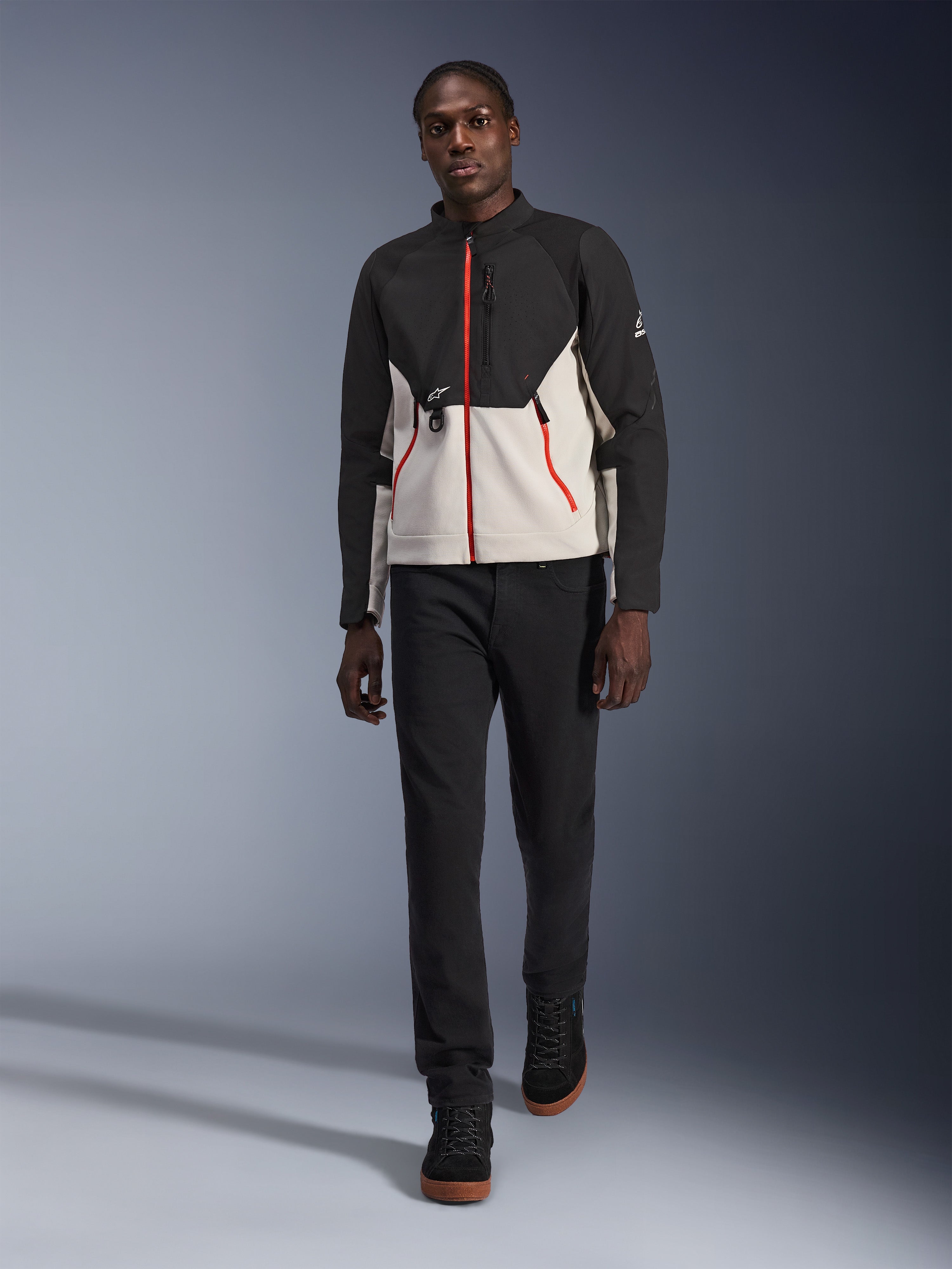 Męski model w kurtce Alpinestars Provoke, miejska kurtka motocyklowa w kolorze czarnym i metalicznym szarym z czerwonymi akcentami przy zamkach, zestawiona z czarnymi spodniami i czarnymi butami za kostkę, nowoczesna odzież ochronna do jazdy miejskiej