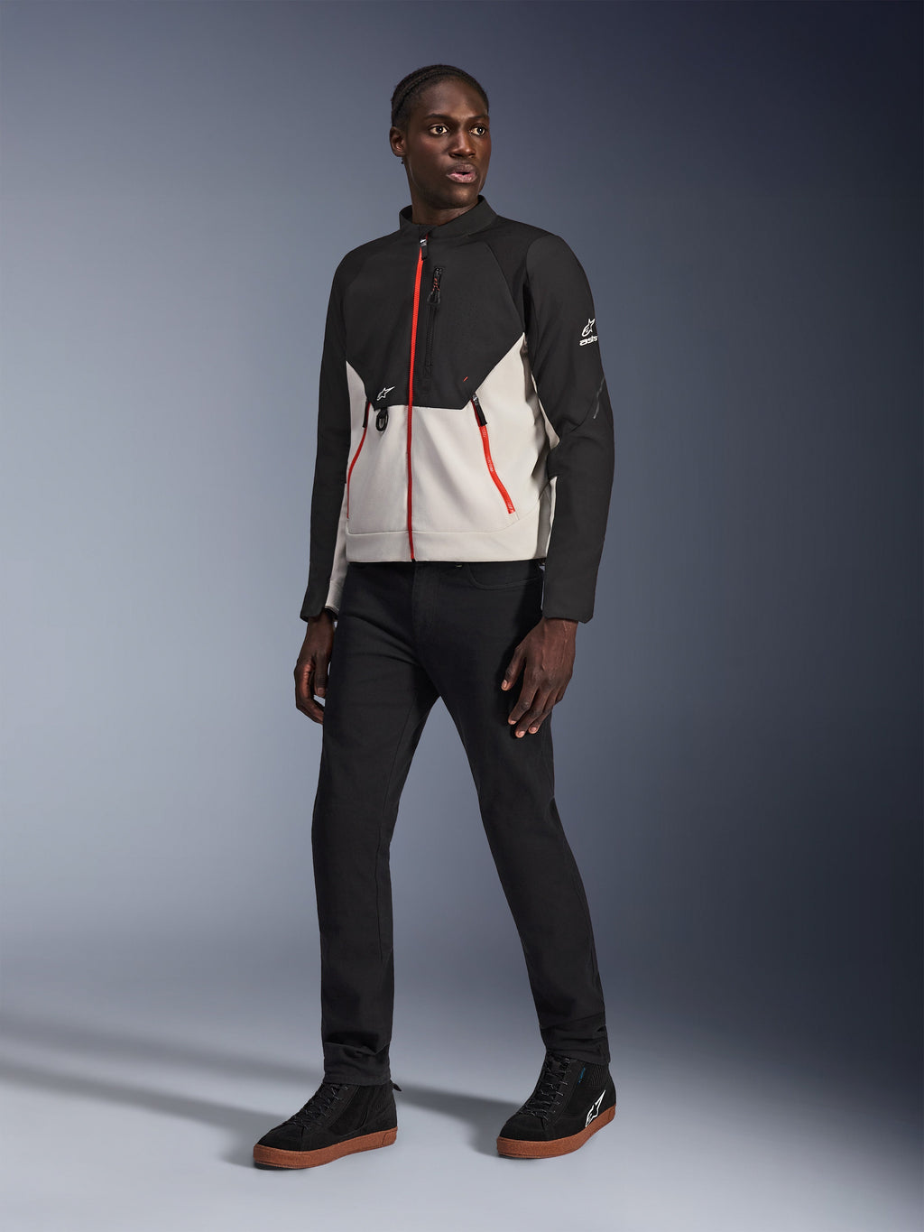 Model ubrany w kurtkę Alpinestars Provoke w kolorze Black Metal Gray z charakterystycznymi czerwonymi zamkami z przodu i przy kieszeniach, czarne spodnie oraz czarne wysokie sneakersy z gumową podeszwą; współczesna miejska odzież motocyklowa w ulicznym stylu.
