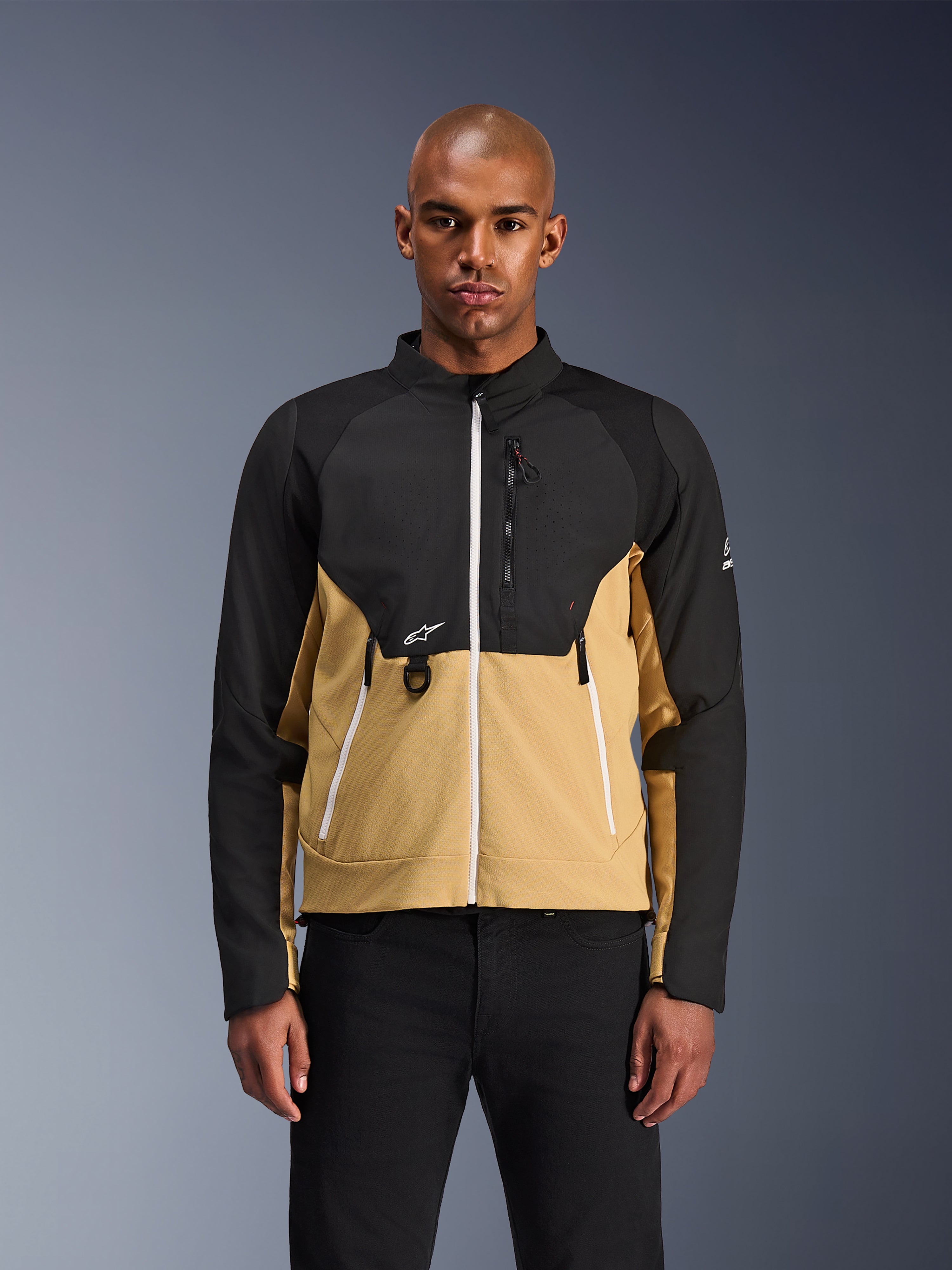 Alpinestars Provoke Jacket, miejska kurtka motocyklowa, czarna i w kolorze camel, widok z przodu na modelu, dwukolorowy design z białymi zamkami, kieszeń na klatce piersiowej z zamkiem i D-ringiem oraz małe białe logo Alpinestars, nowoczesna odzież dla miejskich motocyklistów.