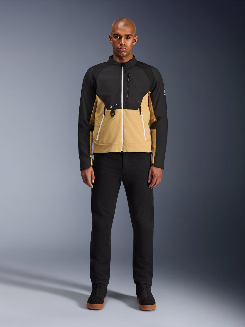 Kurtka Alpinestars Provoke w kolorze Black Camel, miejska kurtka motocyklowa o dwukolorowym czarno-karmelowym designie, prezentowana na modelu w czarnych spodniach i butach, nowoczesna sylwetka z białymi zamkami i techniczną konstrukcją softshell