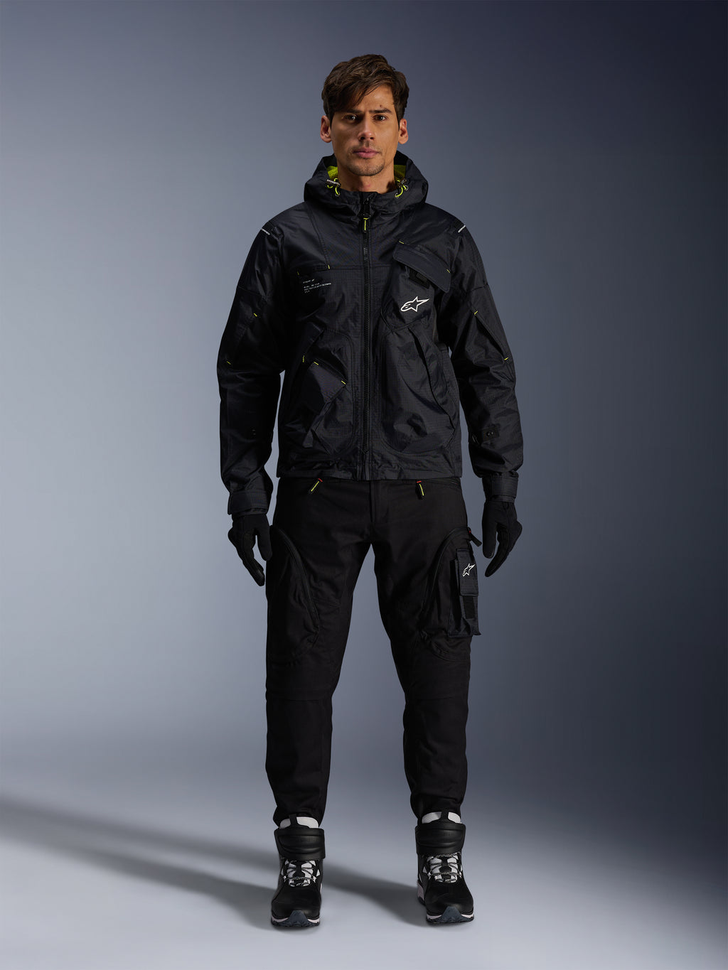 Męski model w kurtce Alpinestars Mogress Air, czarna tekstylna kurtka z kapturem i limonkowymi akcentami, zestawiona z czarnymi spodniami typu utility, czarnymi rękawicami oraz czarno-białymi butami motocyklowymi, miejski design streetwearowy inspirowany motorsportem
