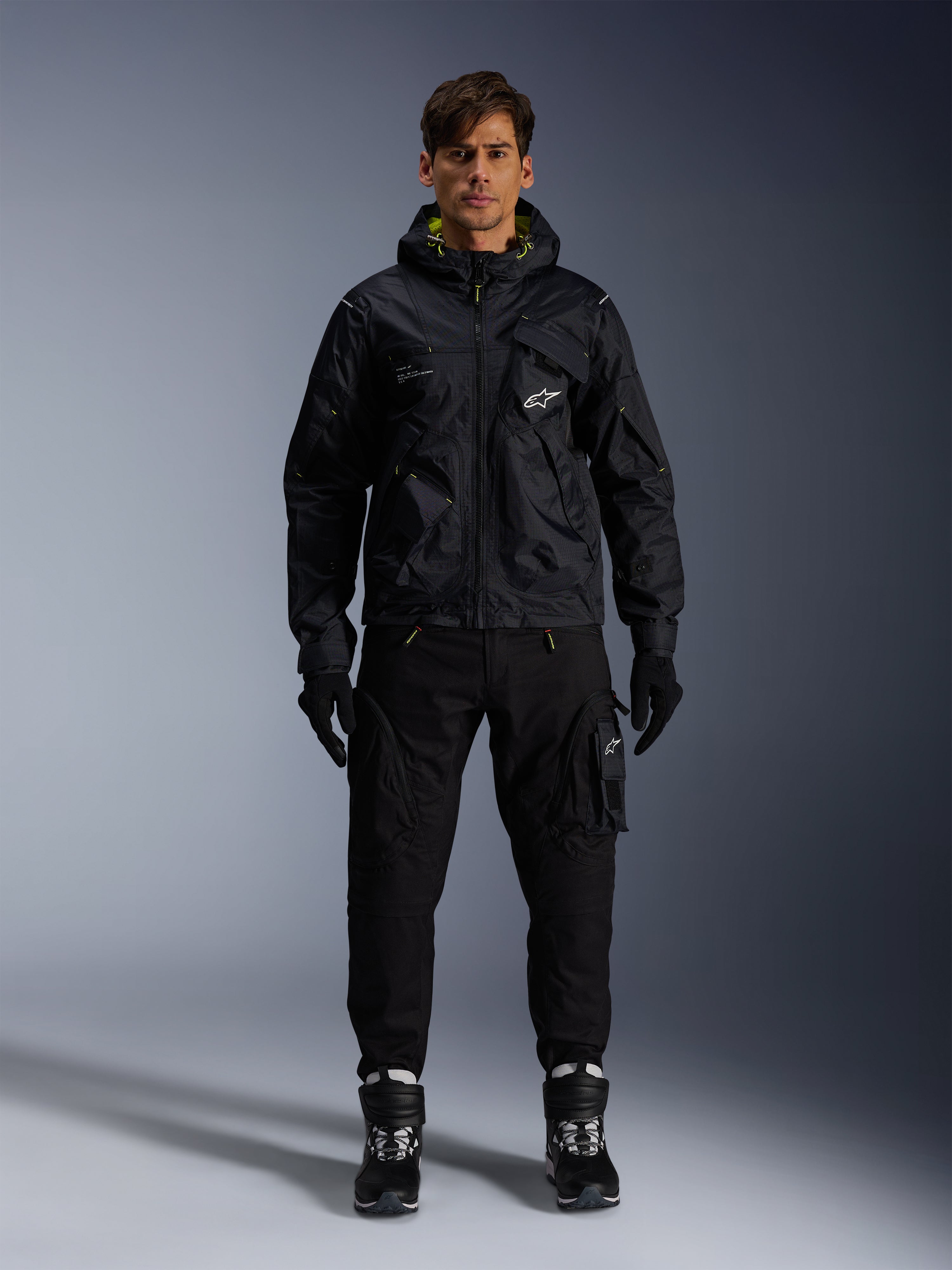 Męski model w kurtce Alpinestars Mogress Air, czarna tekstylna kurtka z kapturem i limonkowymi akcentami, zestawiona z czarnymi spodniami typu utility, czarnymi rękawicami oraz czarno-białymi butami motocyklowymi, miejski design streetwearowy inspirowany motorsportem