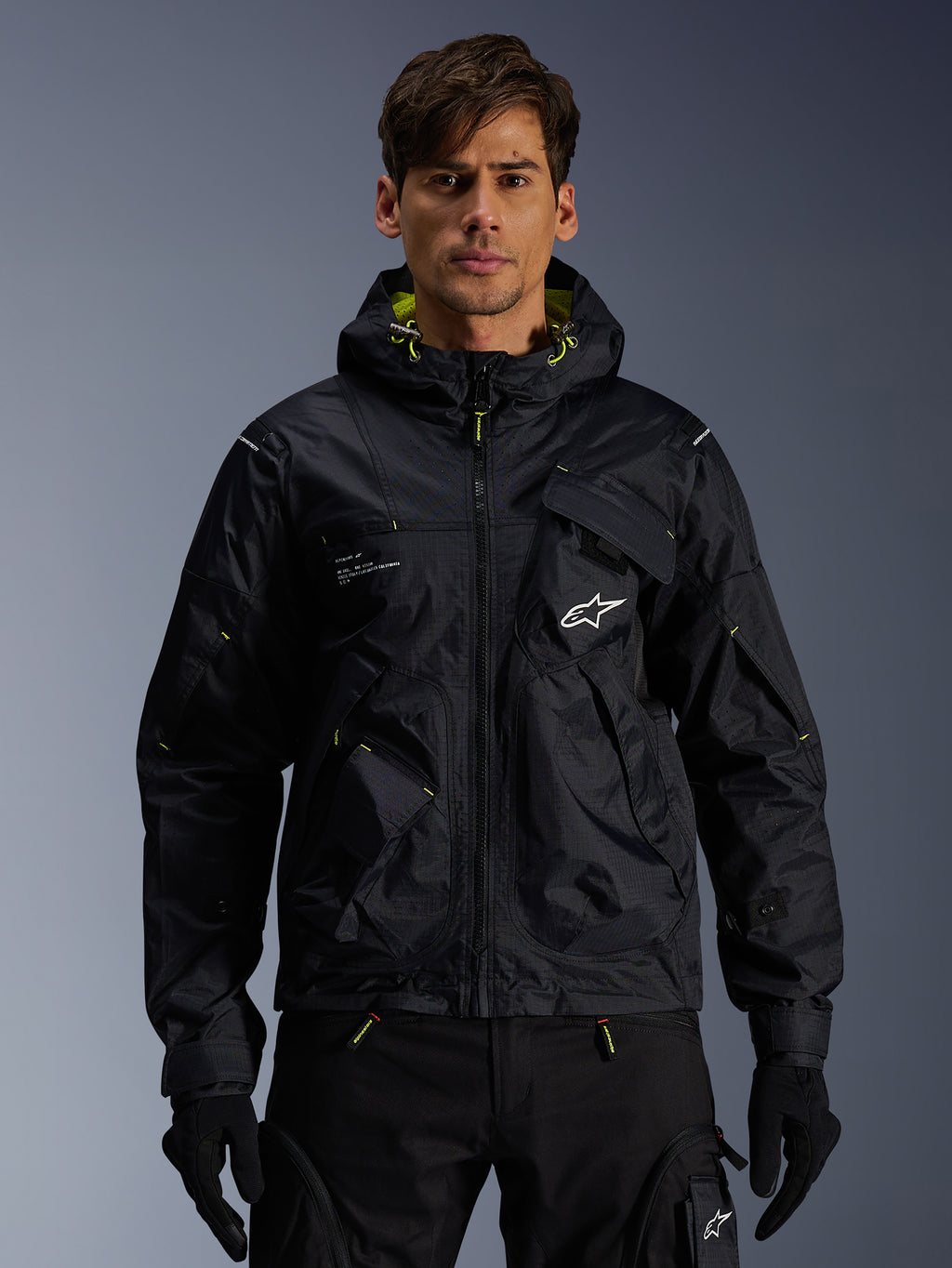 Alpinestars Mogress Air Jacket, miejska kurtka motocyklowa, czarna z limonkowymi akcentami (Lime Punch), prezentowana przez modela w czarnych spodniach i rękawicach, design inspirowany stylem utility z kapturem i białym logo na piersi, lekka odzież tekstylna do jazdy miejskiej