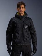Alpinestars Mogress Air Jacket, miejska kurtka motocyklowa, czarna z limonkowymi akcentami (Lime Punch), prezentowana przez modela w czarnych spodniach i rękawicach, design inspirowany stylem utility z kapturem i białym logo na piersi, lekka odzież tekstylna do jazdy miejskiej