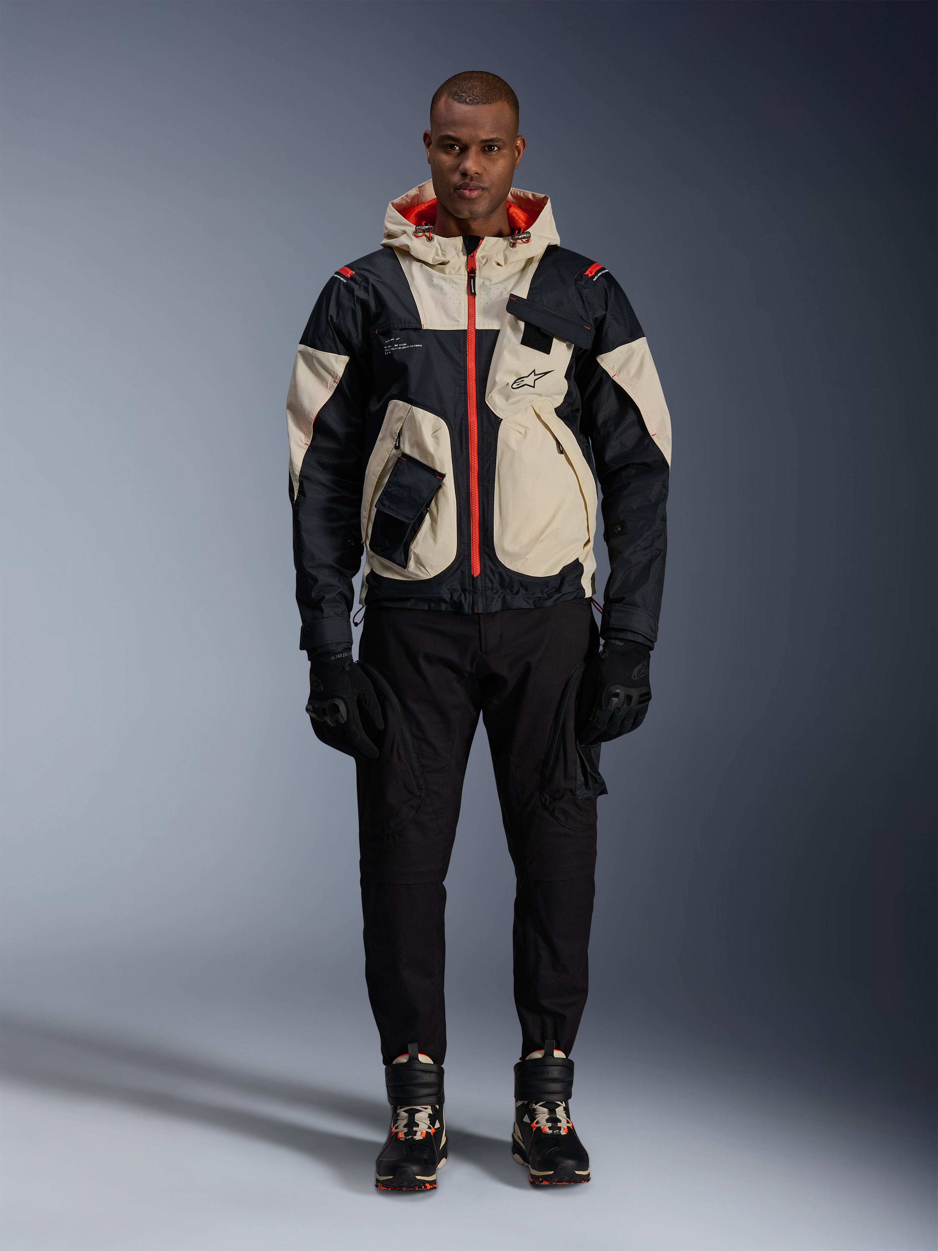 Kurtka Alpinestars Mogress Air, miejska kurtka motocyklowa, kolorystyka Black Almond i Hot Orange, noszona przez modela z czarnymi spodniami i rękawicami, design inspirowany stylem utility z kremowymi panelami i jaskrawopomarańczowymi zamkami, nowoczesny streetwear inspirowany motorsportem.