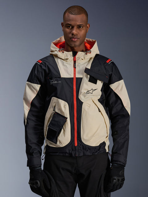 Alpinestars Mogress Air Jacket, miejska kurtka motocyklowa, beżowo-czarna z jaskrawopomarańczowymi zamkami i podszewką kaptura, fason z kapturem i kieszeniami użytkowymi, logo na piersi, noszona z czarnymi rękawicami