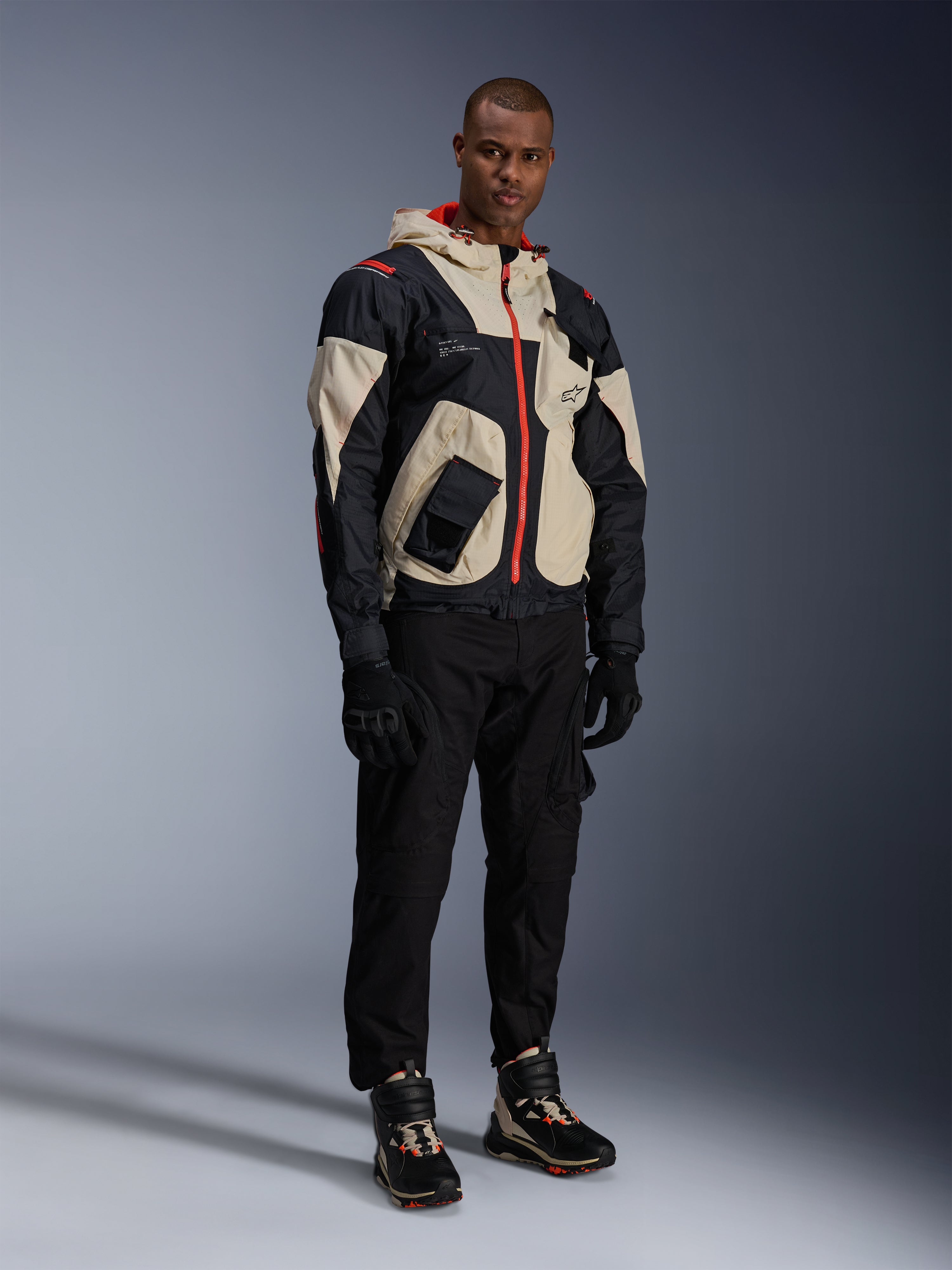 Model ubrany w kurtkę Alpinestars Mogress Air Jacket w kolorach czarnym, beżowym i jaskrawopomarańczowym, miejska kurtka motocyklowa o designie inspirowanym stylem użytkowym, noszona z czarnymi spodniami motocyklowymi, czarnymi rękawicami i dopasowanymi butami technicznymi, współczesny strój motocyklowy w stylu ulicznym