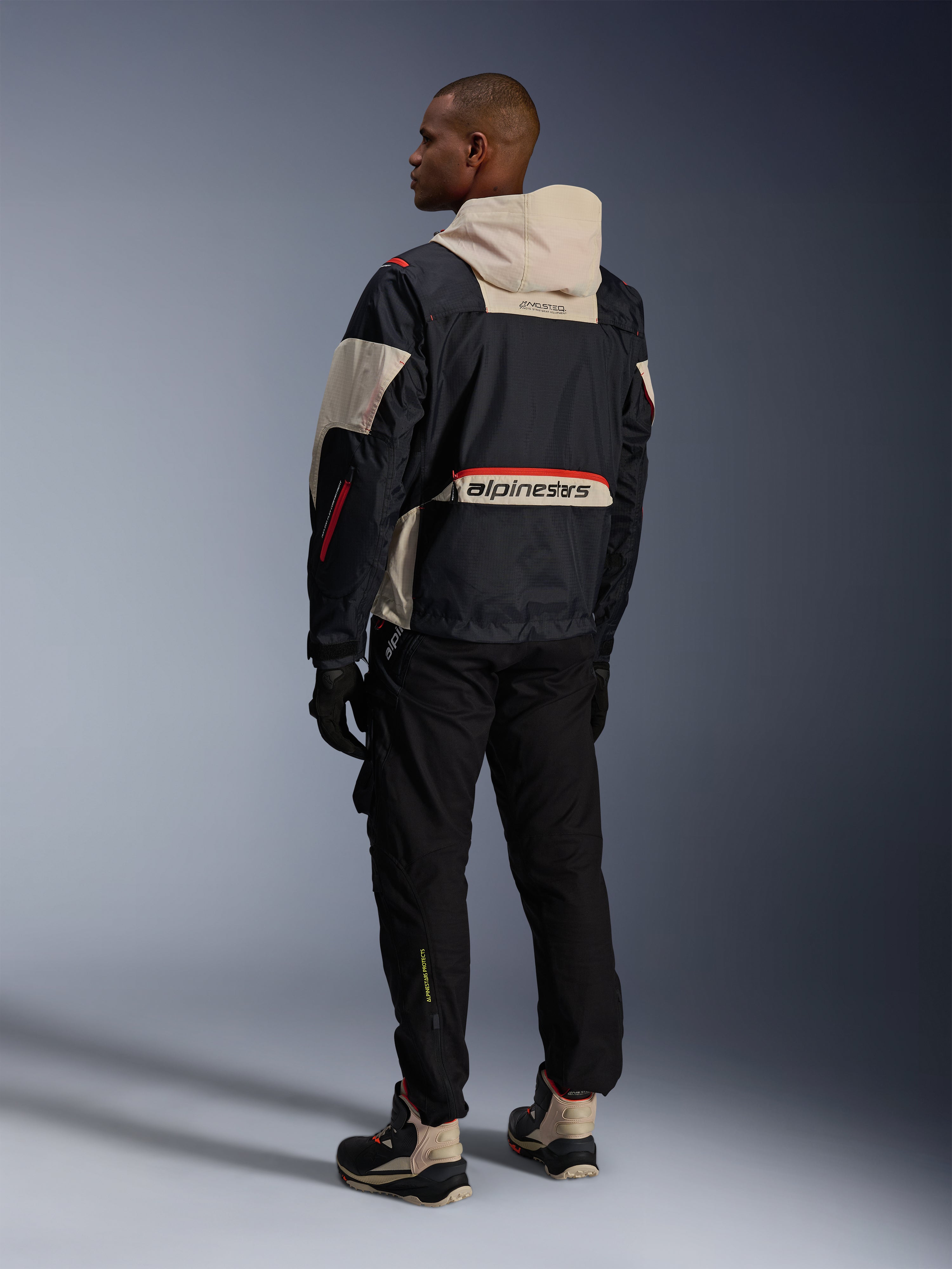 Alpinestars Mogress Air Jacket, Black Almond Hot Orange, widok od tyłu motocyklisty w miejskim stroju motocyklowym, kaptur i dolny panel na plecach w kolorze migdałowym z dużym logo Alpinestars, pomarańczowe detale zamków, w zestawie z czarnymi spodniami i wysokimi butami motocyklowymi.