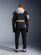 Widok z tyłu motocyklisty w kurtce Alpinestars Mogress Air Jacket w kolorach Black, Almond i Hot Orange, z kremowym kapturem, dużym logo Alpinestars na tylnej kieszeni i pomarańczowymi akcentami zamków, w zestawieniu z czarnymi spodniami i kremowymi butami, miejski styl motocyklowy.