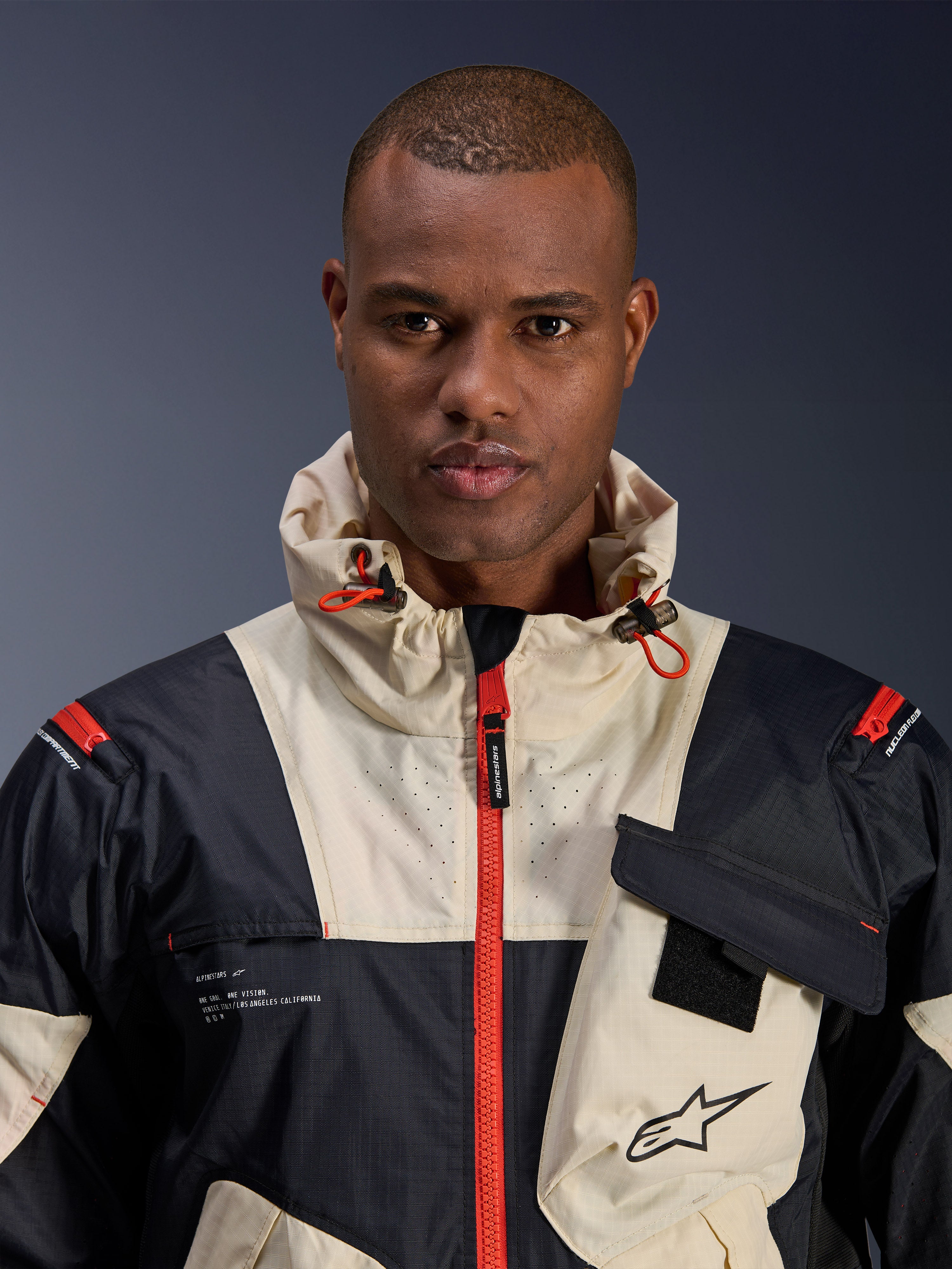 Model w kurtce Alpinestars Mogress Air, miejska kurtka motocyklowa, kolory Black Almond i Hot Orange, zbliżenie prezentujące design color-block, teksturę ripstop, kołnierz z kapturem z pomarańczowymi ściągaczami oraz detale przedniego zamka