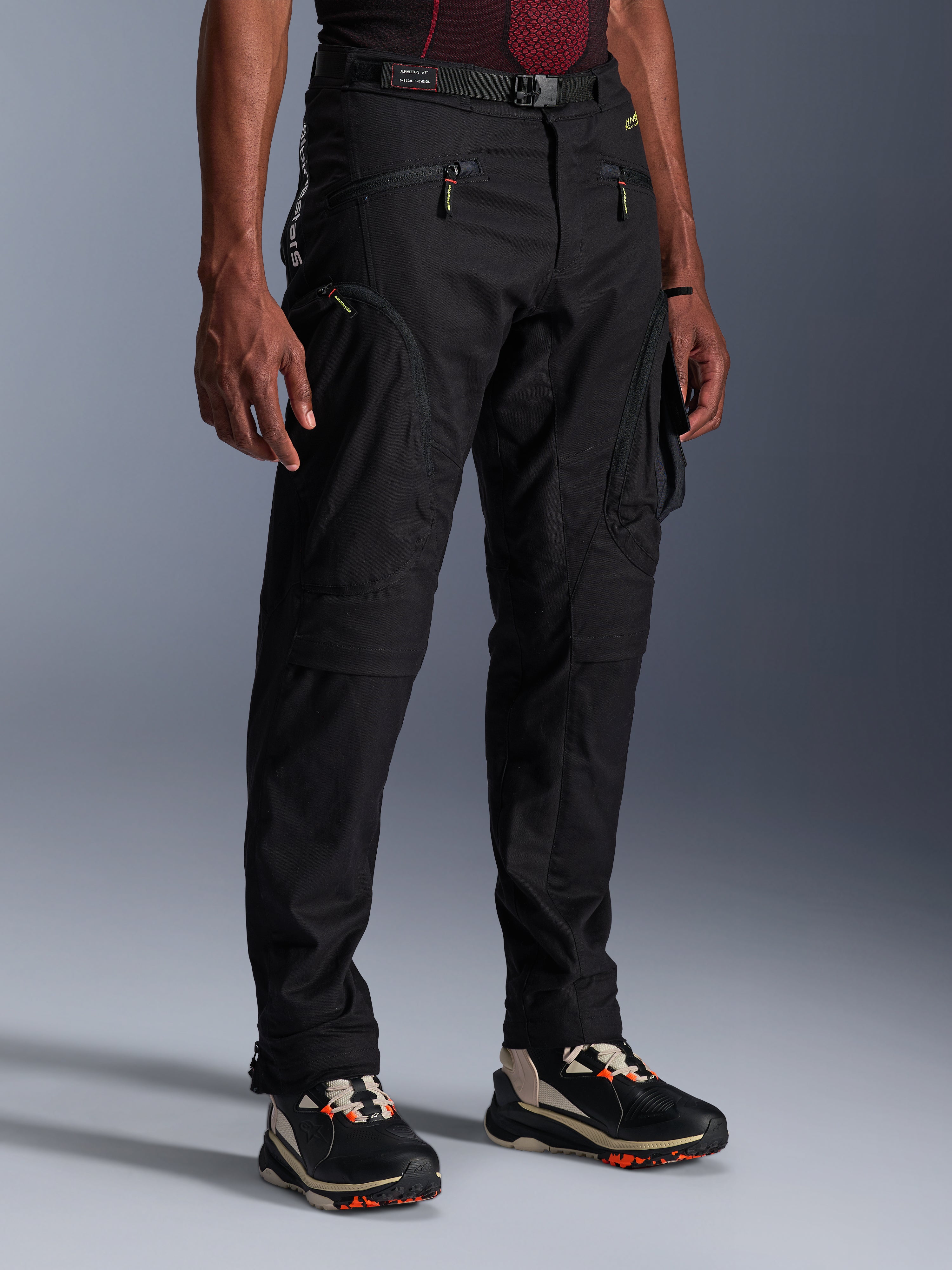 Alpinestars Acteon Pants, czarne spodnie motocyklowe prezentowane na modelu, o nowoczesnym, zwężanym kroju, z kieszeniami cargo zapinanymi na zamek, regulowanym paskiem w talii i technicznymi sneakersami, miejska odzież motocyklowa