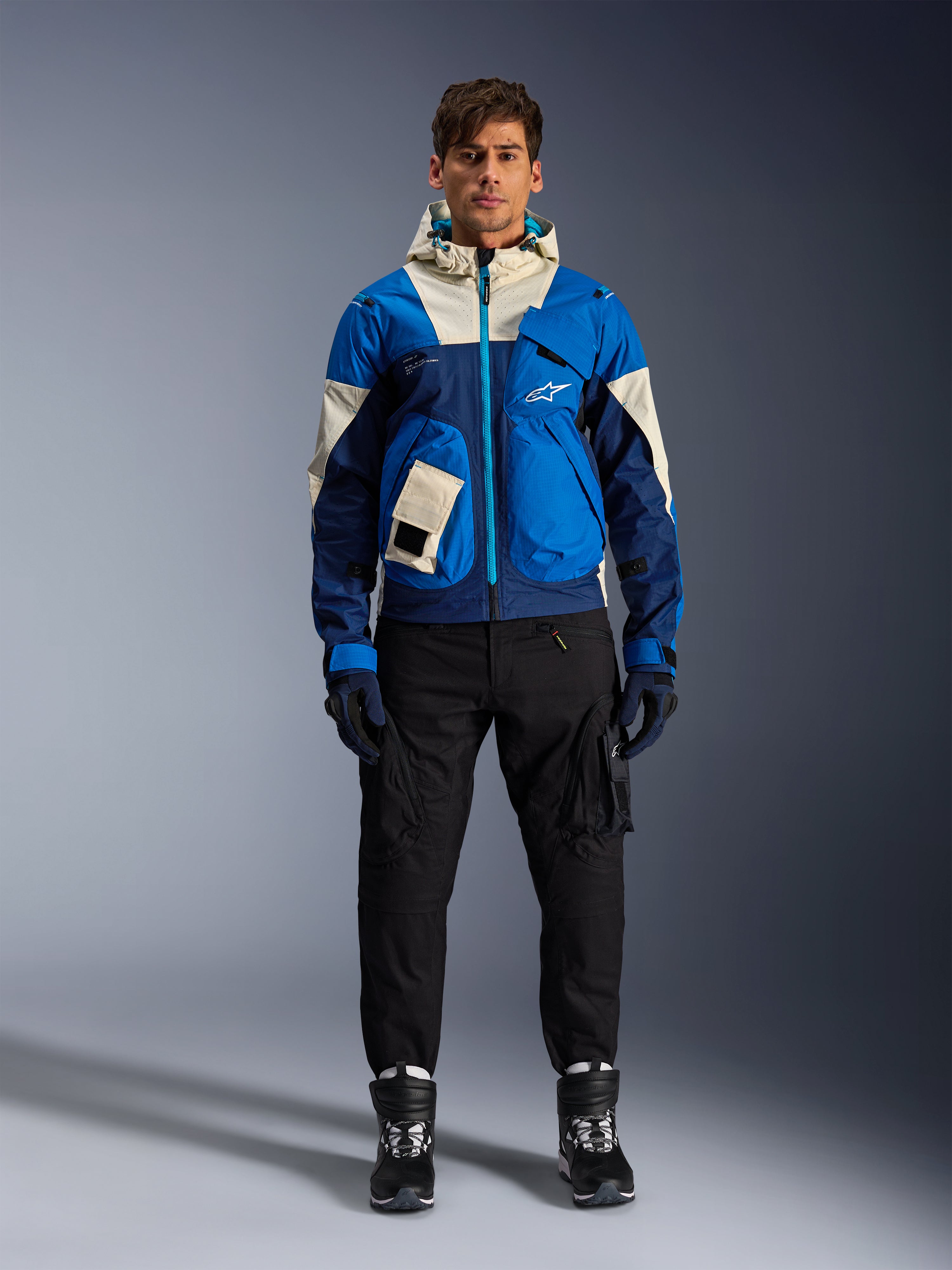 Kurtka Alpinestars Mogress Air, miejska kurtka motocyklowa Blue Almond, noszona z czarnymi spodniami i butami, o technicznym designie w niebieskie i kremowe bloki kolorystyczne, z funkcjonalnymi kieszeniami i kapturem, nowoczesna odzież motocyklowa inspirowana stylem streetwear