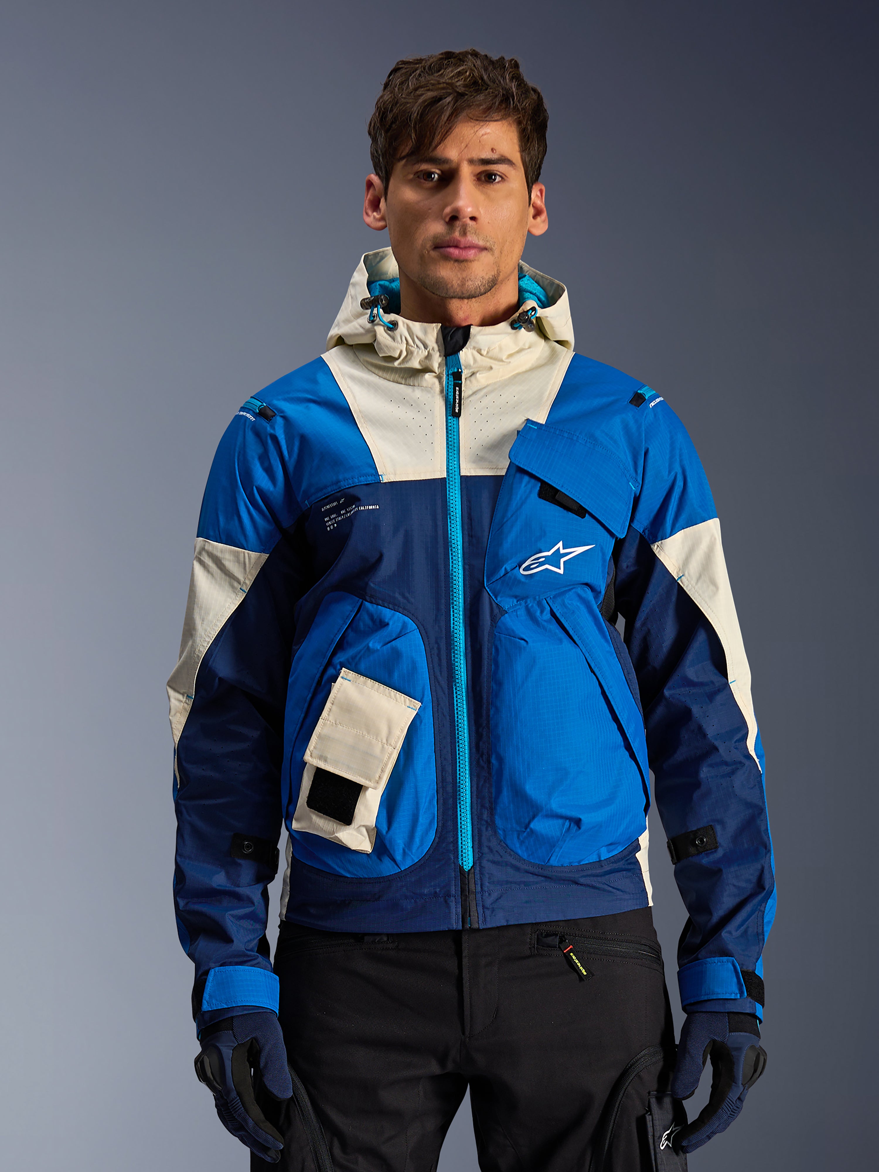 Alpinestars Mogress Air Jacket w kolorze Blue Almond, miejska kurtka motocyklowa z kapturem z kontrastującymi niebieskimi i białawymi panelami, kieszeniami użytkowymi i morskim przednim zamkiem błyskawicznym, noszona przez modela w czarnych spodniach i rękawicach