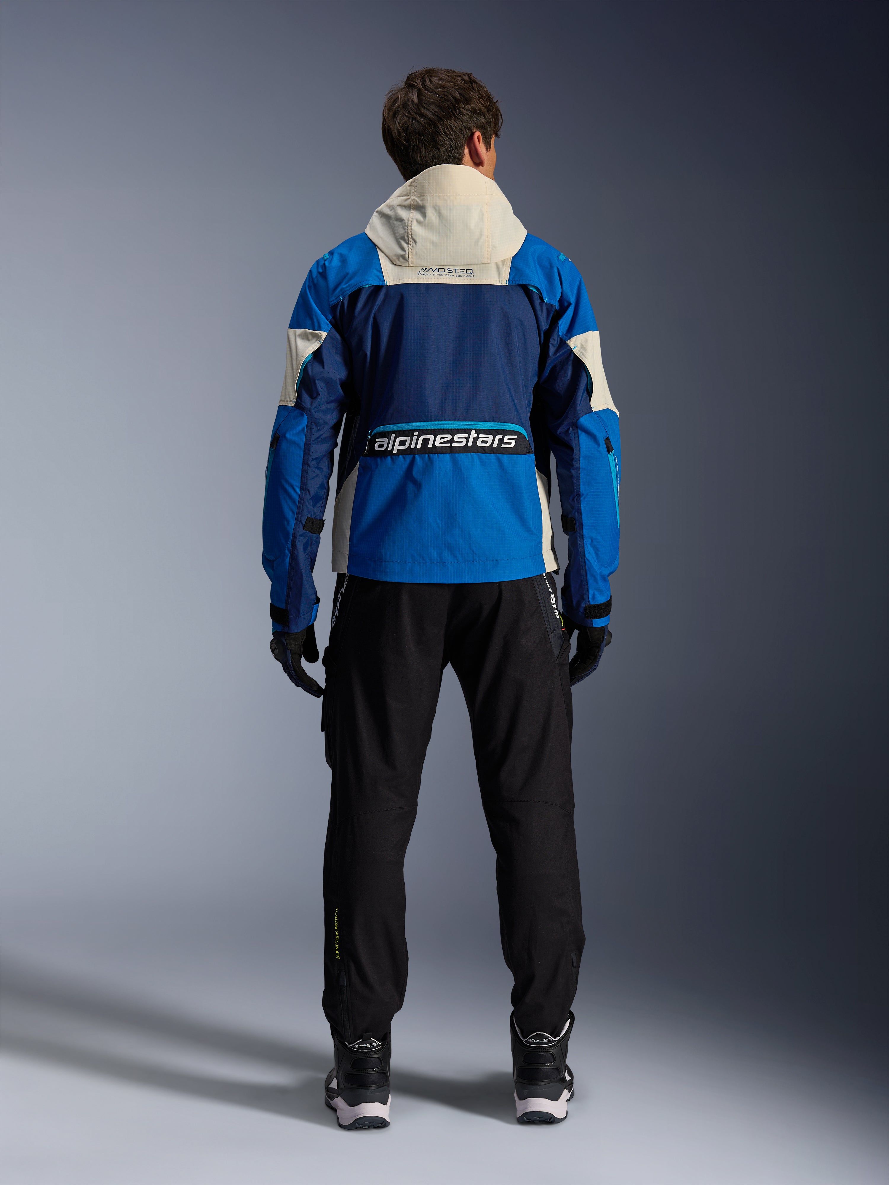 Kurtka Alpinestars Mogress Air w kolorze Blue Almond, widok od tyłu motocyklisty w kurtce motocyklowej, granatowe i jasnoniebieskie panele z kapturem w kolorze złamanej bieli, wyraźne logo Alpinestars w dolnej części pleców, nowoczesny miejski styl odzieży motocyklowej.