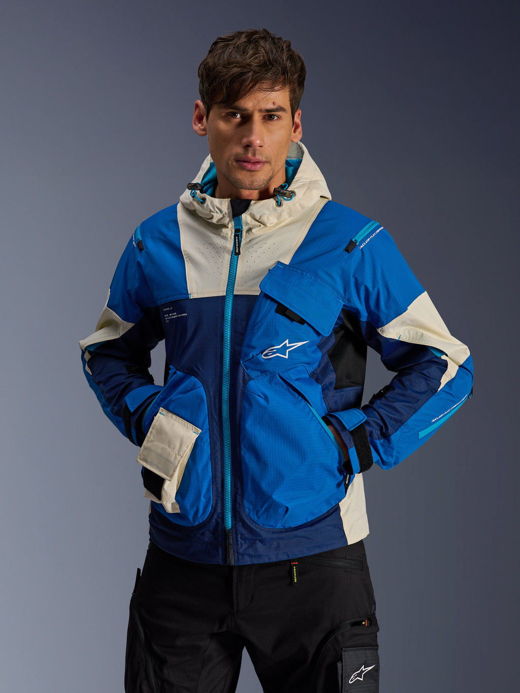 Męski model w kurtce Alpinestars Mogress Air w kolorze Blue Almond, miejska kurtka motocyklowa z jaskrawoniebieskimi i granatowymi panelami kolorystycznymi, kremowe akcenty na kapturze i kieszeniach, techniczna tkanina ripstop z kieszeniami typu utility, nowoczesny streetwear inspirowany motorsportem