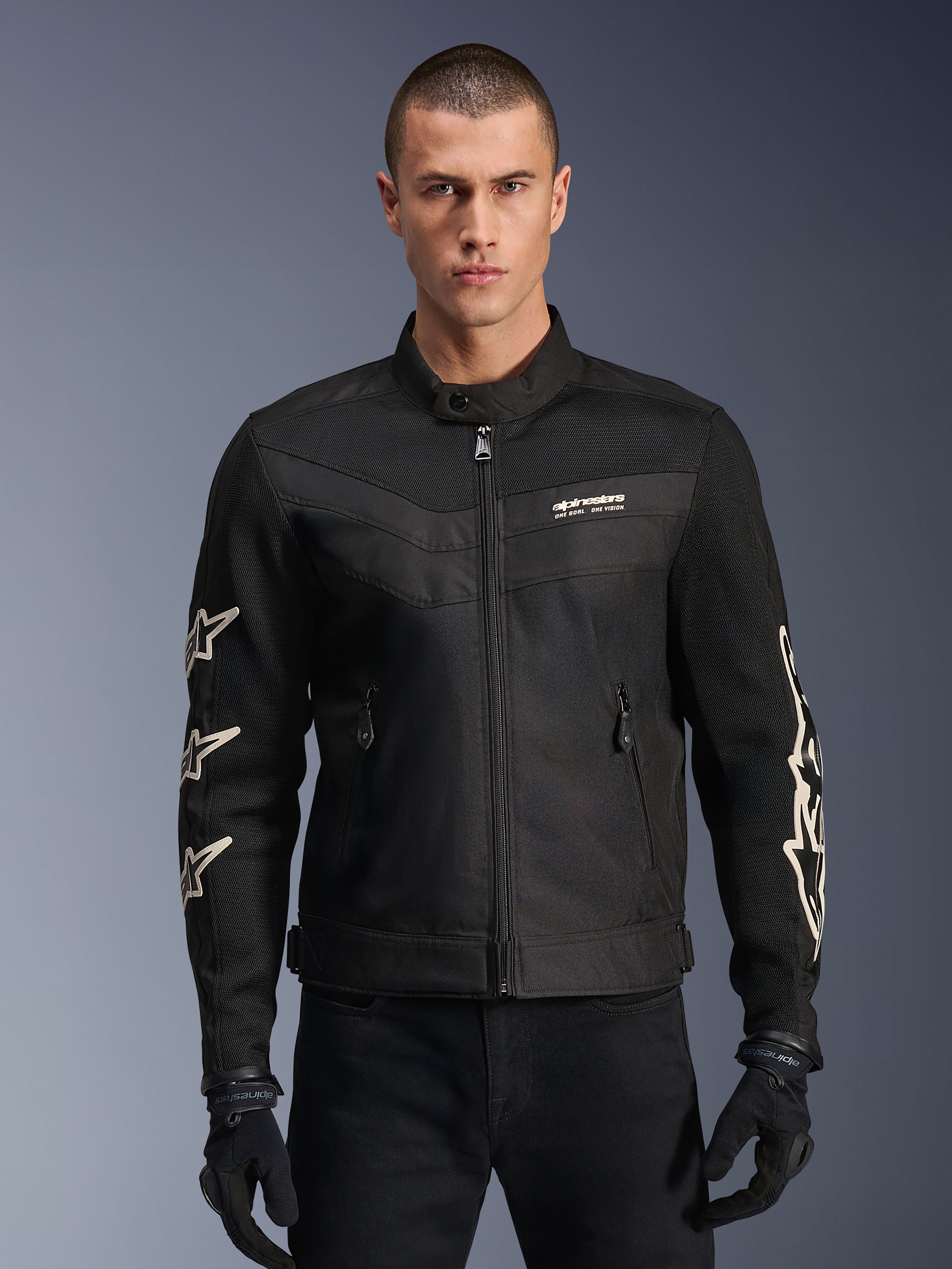 Alpinestars T-Dyno Air Jacket, czarna tekstylna kurtka motocyklowa, z białymi grafikami gwiazd na rękawach i logo na klatce piersiowej, panele z siatki zapewniające wentylację, krój motocyklowy inspirowany stylem vintage, noszona z czarnymi rękawicami i spodniami