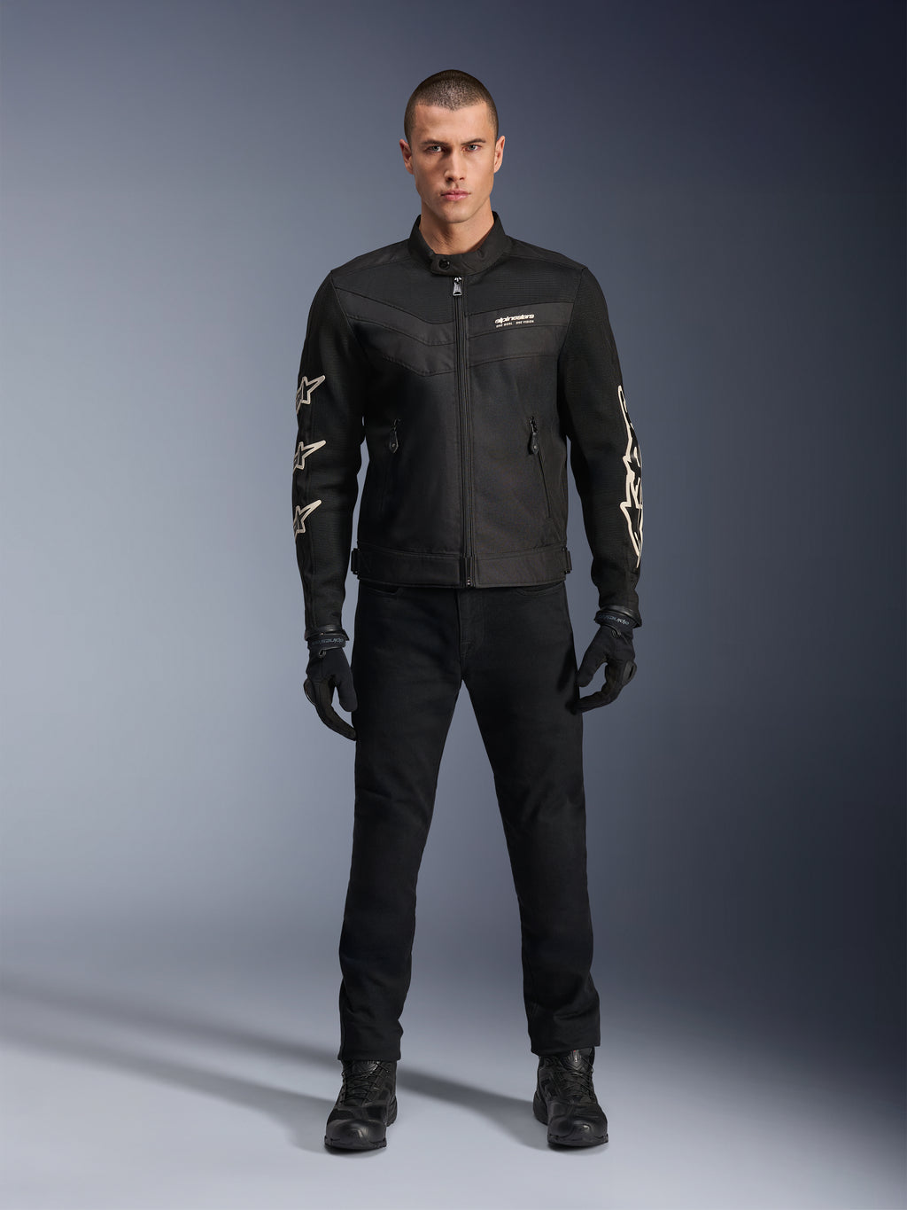 Model w kurtce Alpinestars T-Dyno Air, czarna, lekka motocyklowa kurtka tekstylna z oddychającą siatką 3D, białe logo gwiazdy na rękawach, zestawiona z czarnymi spodniami, rękawicami i butami, widok z przodu